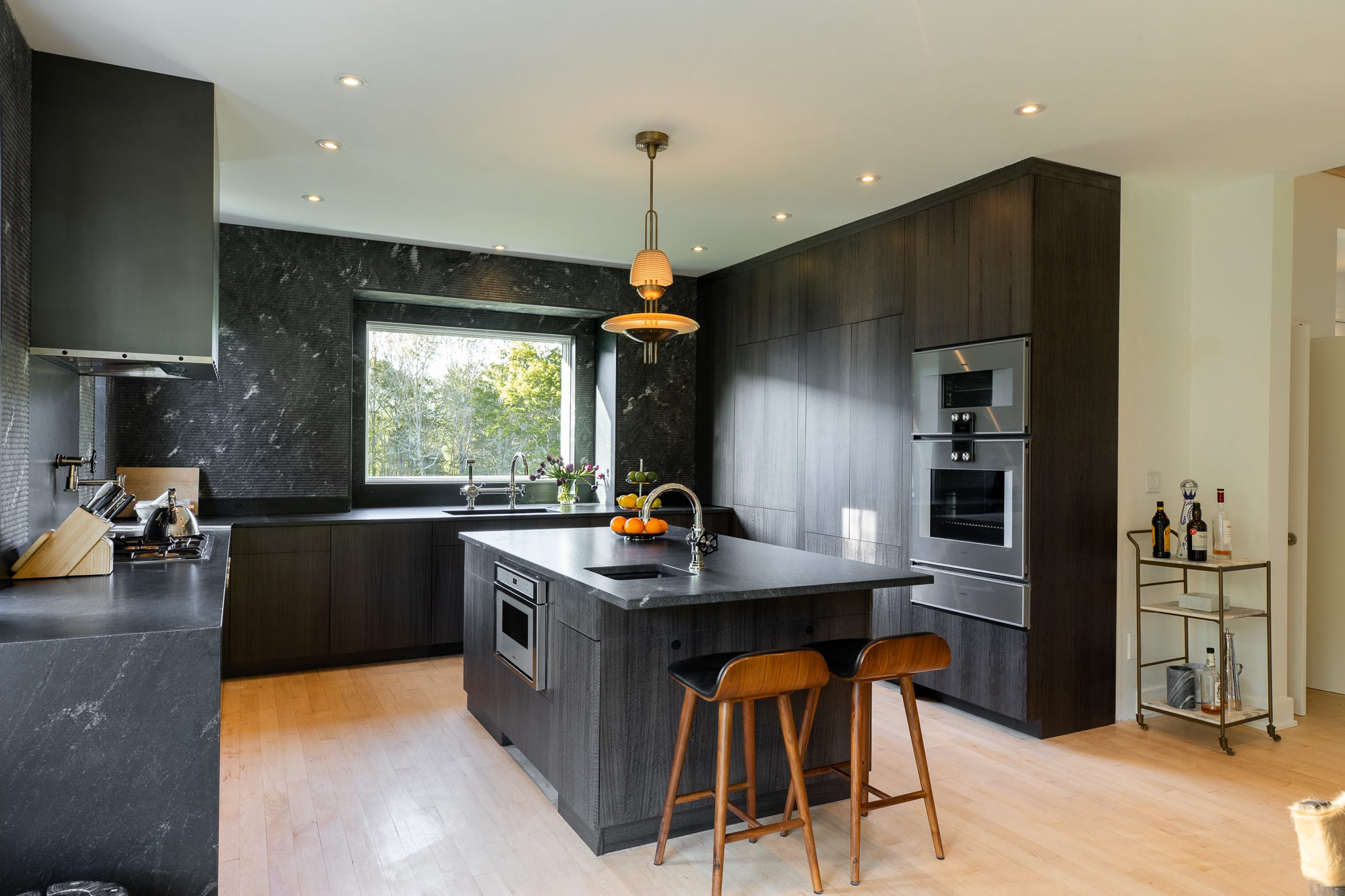 Modern kitchen featuring dark wood cabinetry, a large island with a sink and two bar stools, stainless steel appliances, and a dark stone backsplash and countertops. A large window overlooks greenery.