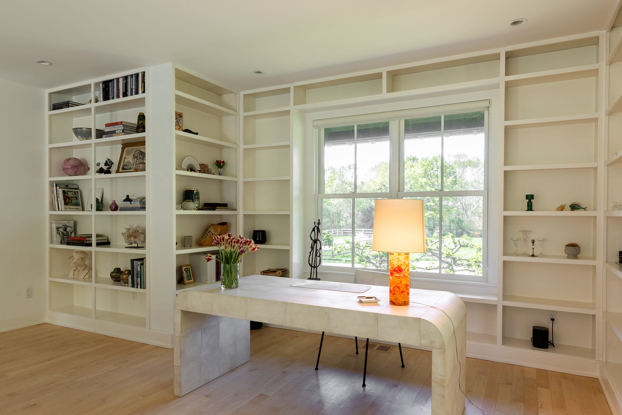 Bright office space with light wood floors and white walls, featuring extensive built-in white bookshelves. A modern white desk with a unique orange lamp is positioned in front of a large window overlooking the outdoors.
