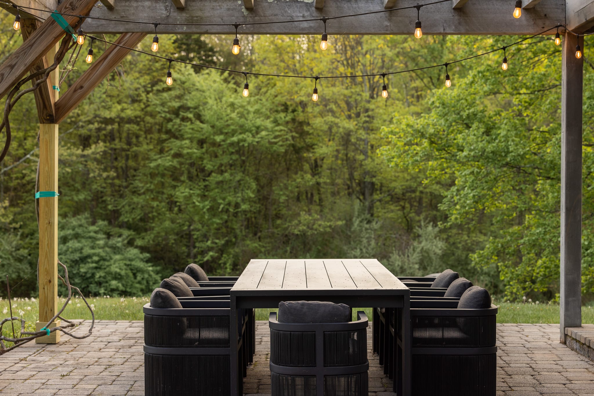 A close-up of an outdoor dining table, made of dark wood or metal, surrounded by black woven chairs with dark cushions, all situated on a paver patio under a wooden pergola with string lights. The background reveals a lush green forest under a bright sky.