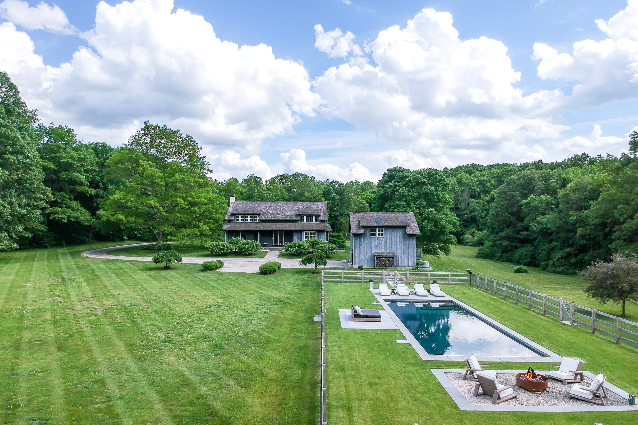 Aerial view of a large gray house with a shingled roof, situated on a sprawling green lawn. A rectangular swimming pool with a fire pit area is visible to the right, enclosed by a wooden fence, surrounded by mature trees.