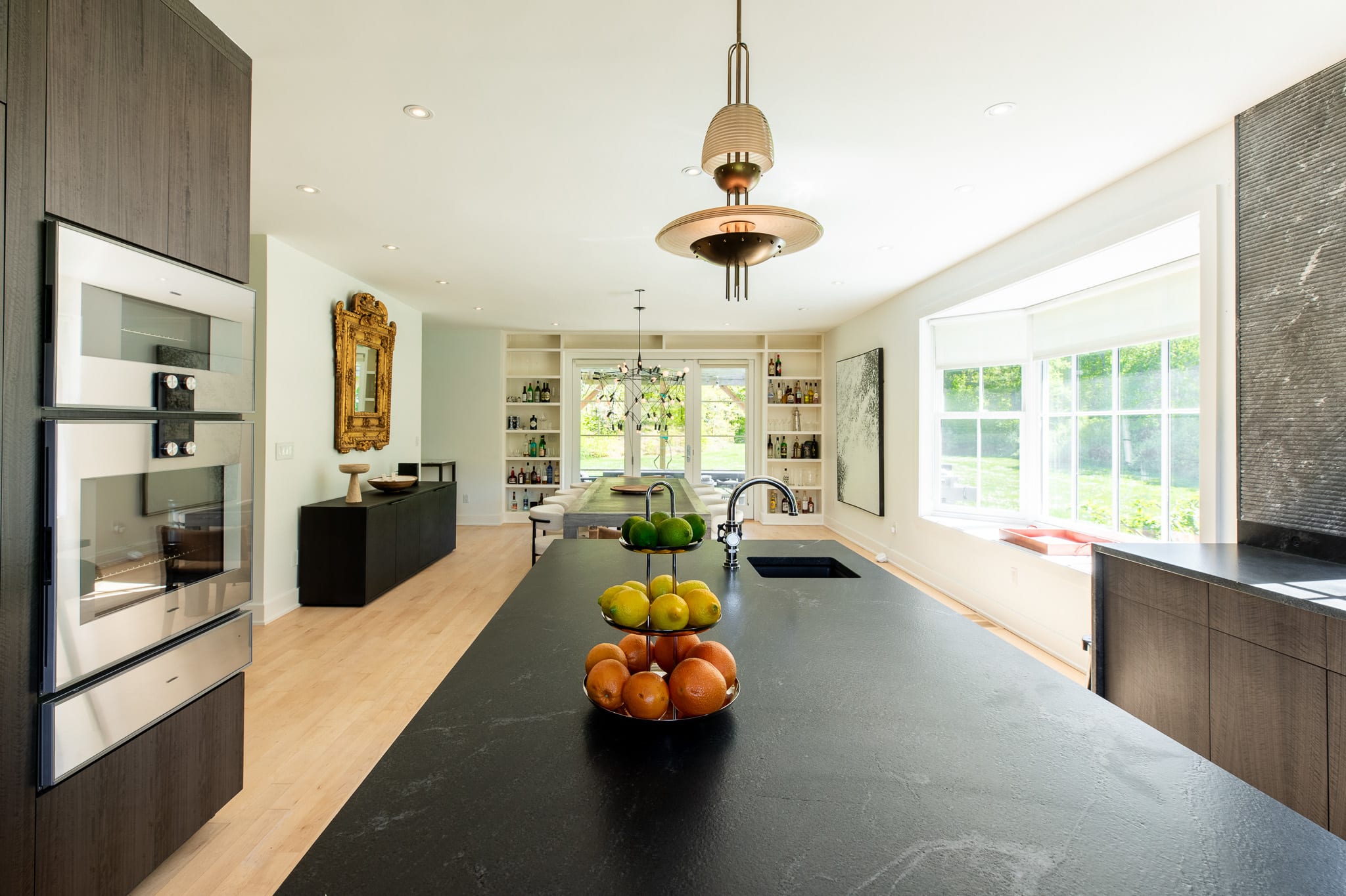 View from the kitchen island looking into an open-concept dining area. The kitchen features dark stone countertops, a sink, and a fruit bowl, with built-in ovens visible on the left.
