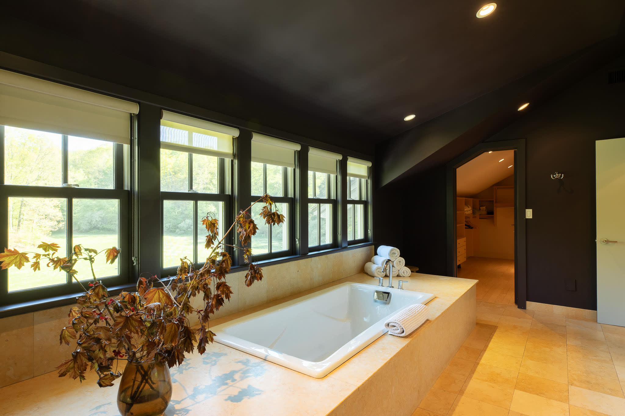 Built-in bathtub with light tile surround in a dark-walled bathroom, offering views of the outdoors through a row of windows. A doorway leads to another room.