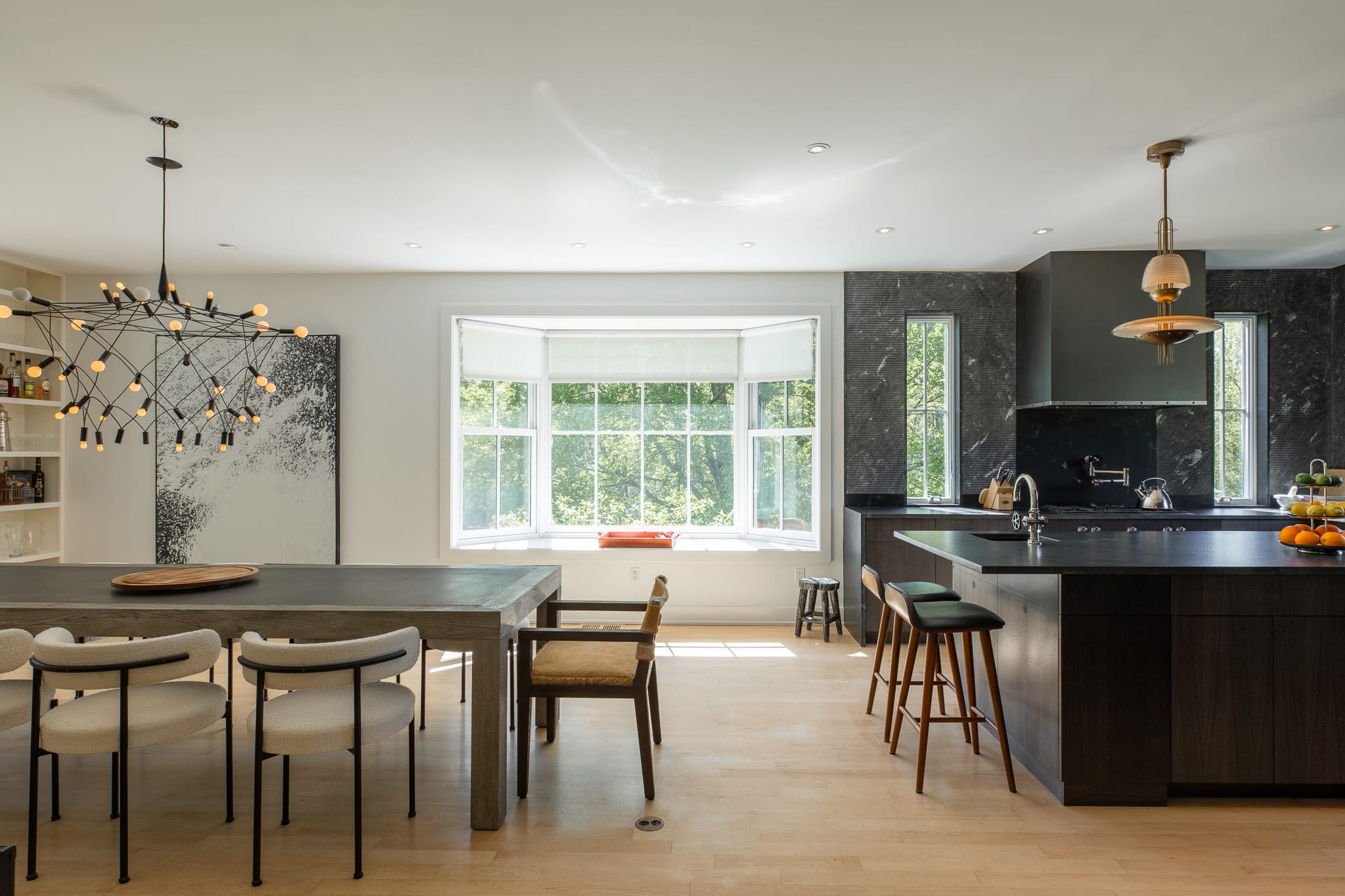 Open-concept dining area with a long dark wood table and modern chairs, adjacent to the kitchen. A large bay window offers views of the outdoors, and a contemporary chandelier hangs above the table.