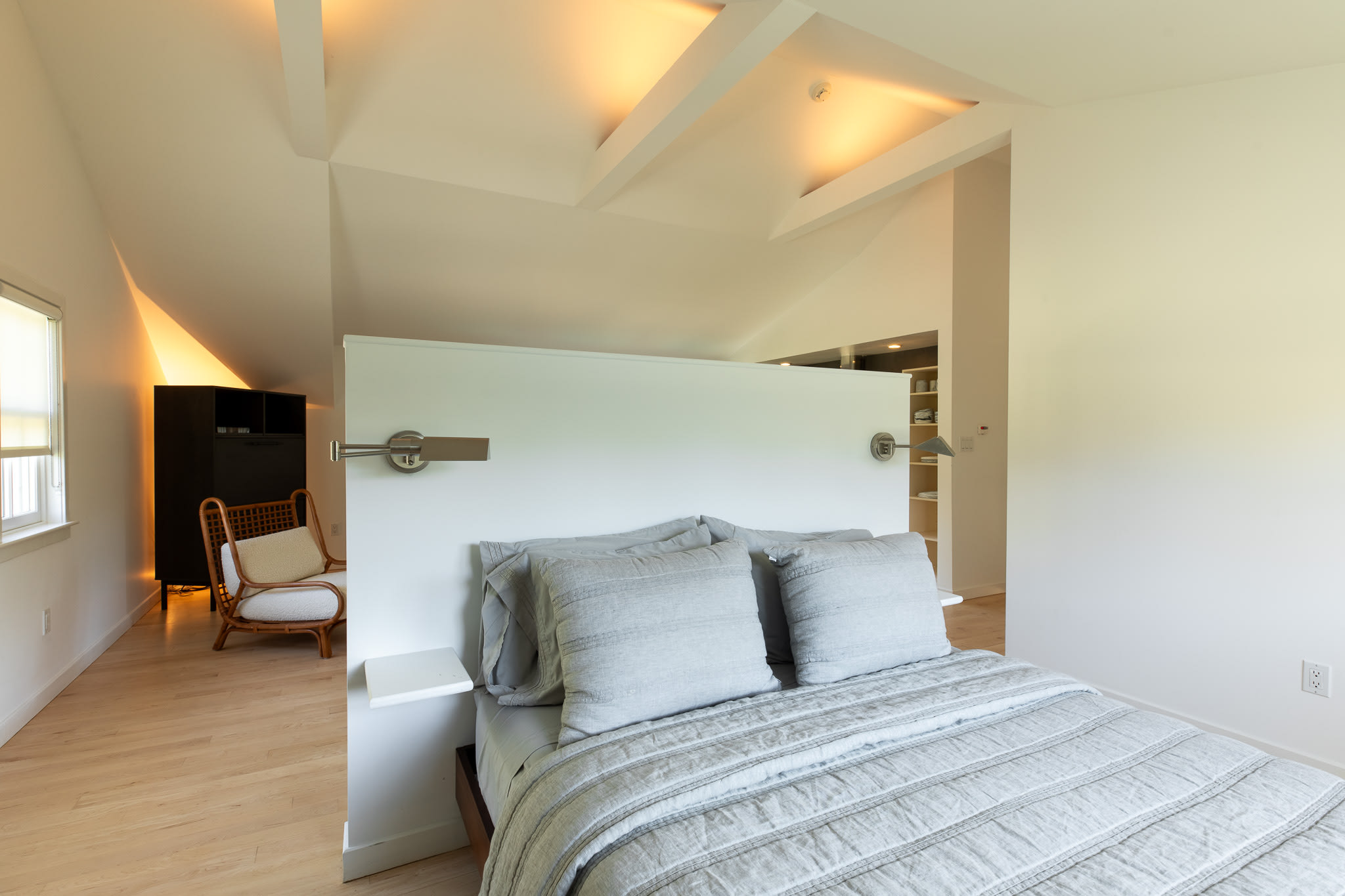 Bright bedroom area featuring a large bed with light grey bedding and pillows, positioned behind a white partition wall, with a vaulted ceiling and warm accent lighting.