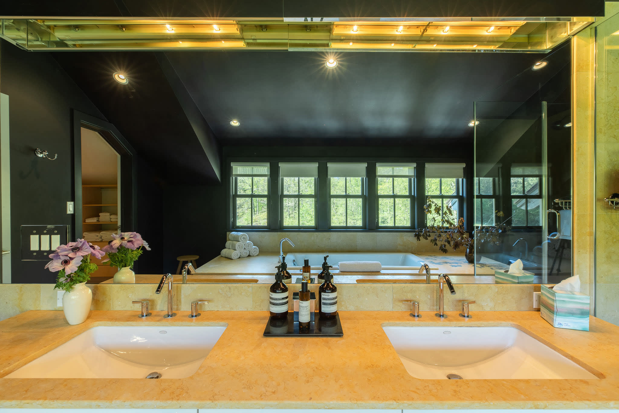Close-up of the double vanity with two sinks, light countertops, and a large mirror reflecting the bathtub and windows in the dark-walled bathroom.