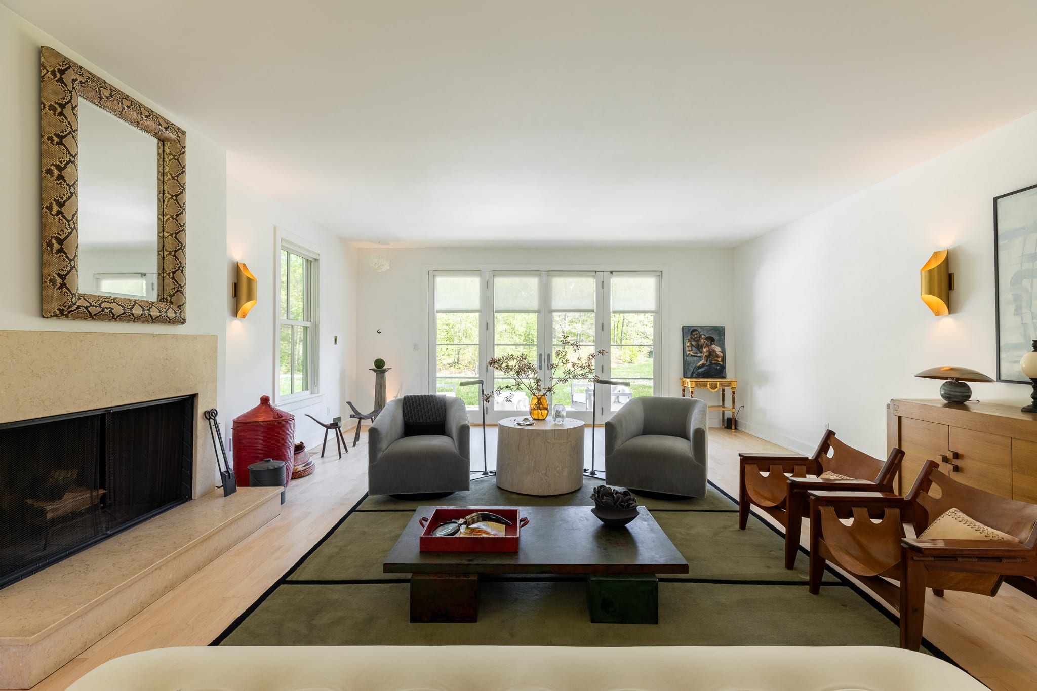 The living room features a stone fireplace with a large ornate mirror above, flanked by golden wall sconces. Two gray armchairs and two leather sling chairs are arranged around a dark coffee table on a green rug, with French doors in the background.