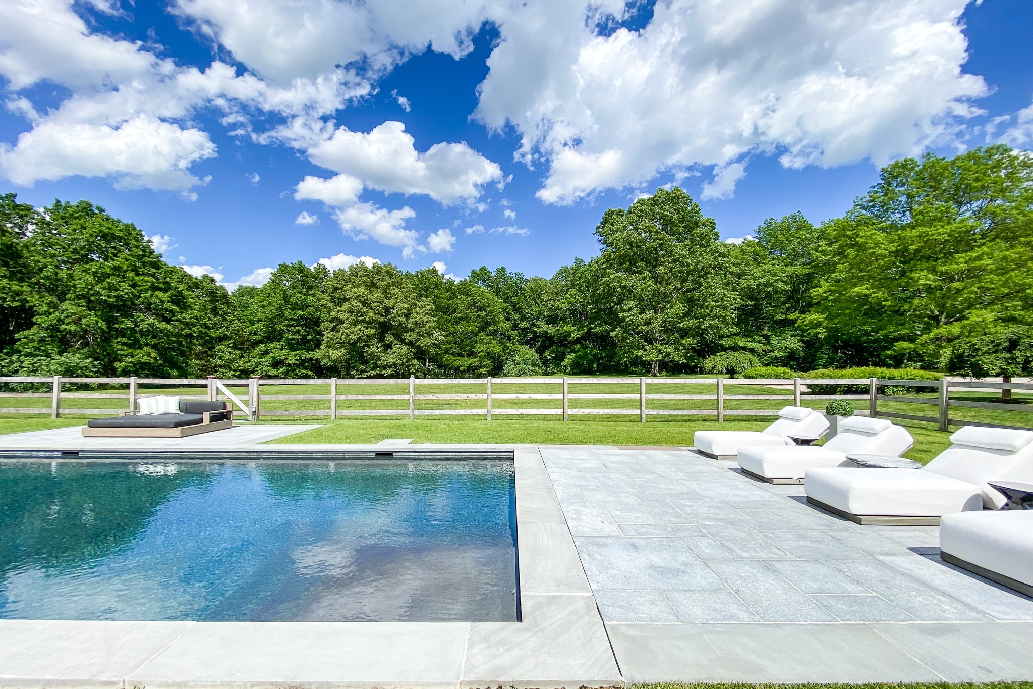 Serene rectangular swimming pool with dark water, bordered by light gray pavers and several white lounge chairs. A wooden fence separates the pool area from a lush green lawn and a dense backdrop of trees under a partly cloudy sky.