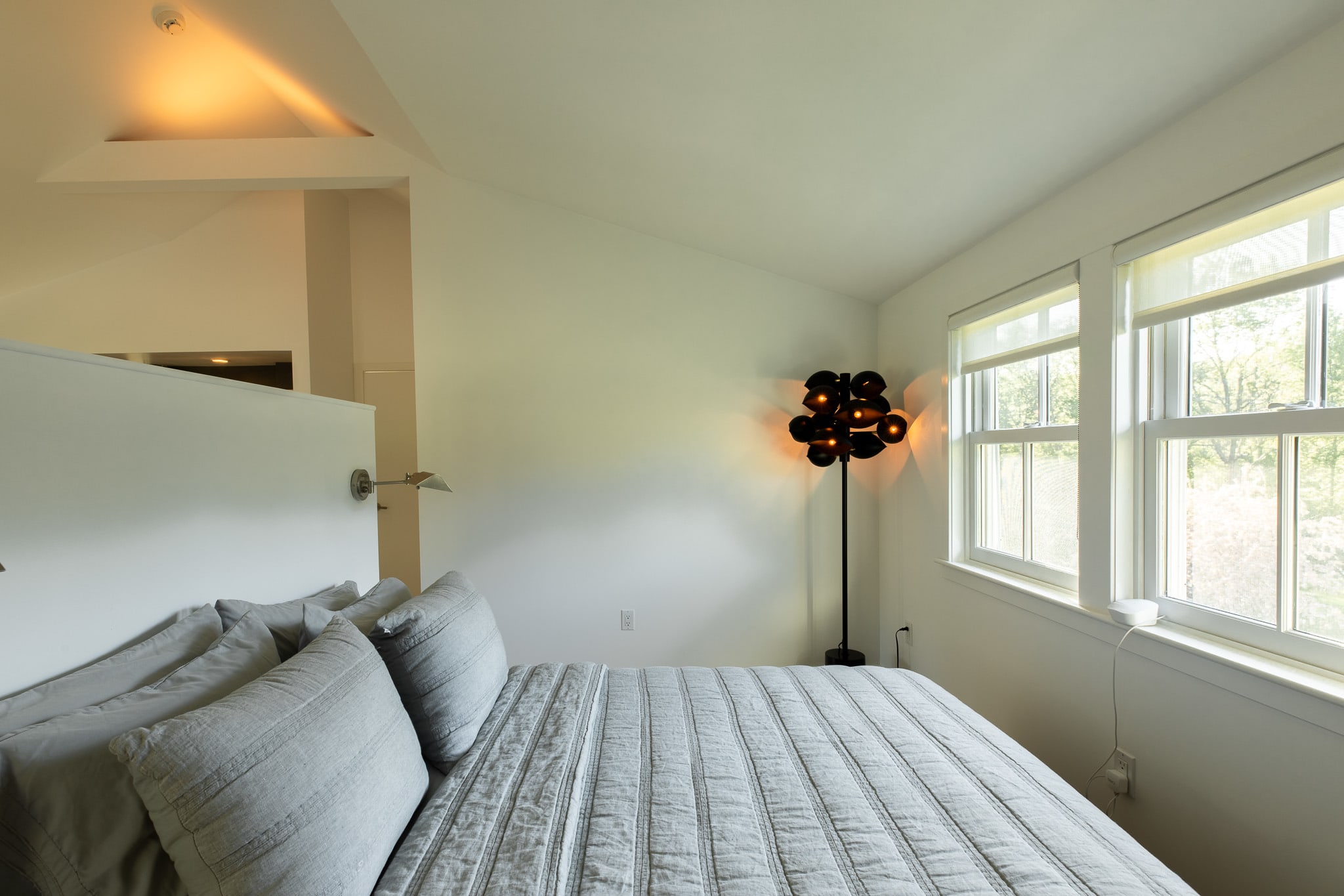 Serene bedroom area with a large bed, light grey bedding, and a unique black floor lamp, illuminated by natural light from two windows and warm ceiling lights.