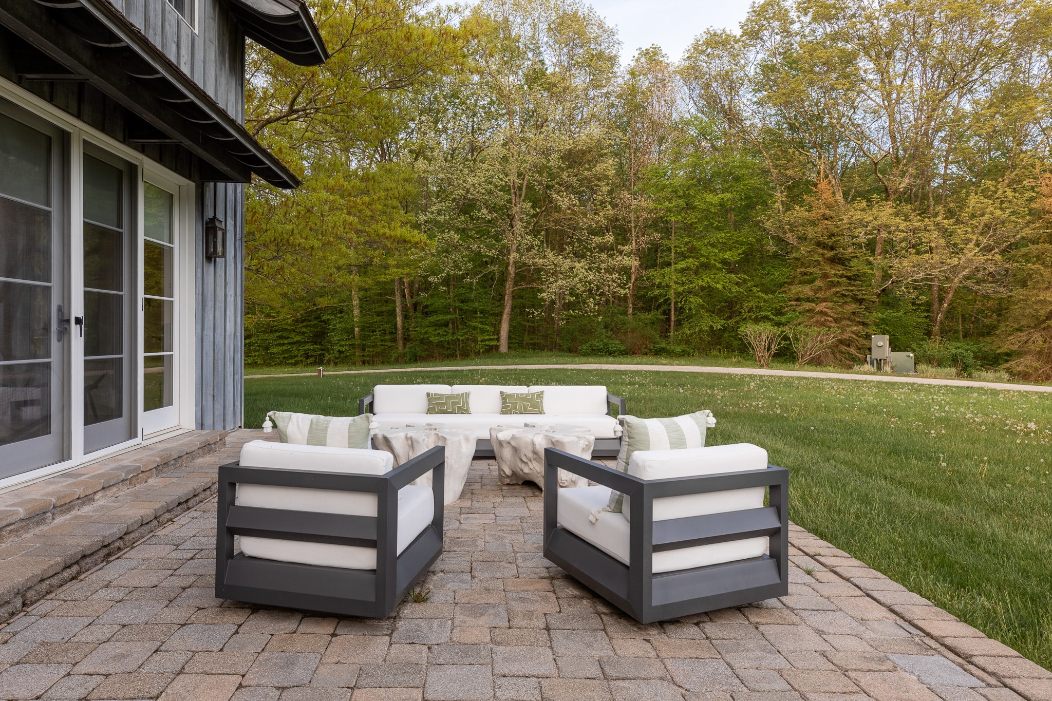 Outdoor patio area with gray paver stones, featuring a white sofa and two armchairs with dark frames. The patio is adjacent to the house, overlooking a green lawn and a dense backdrop of trees.