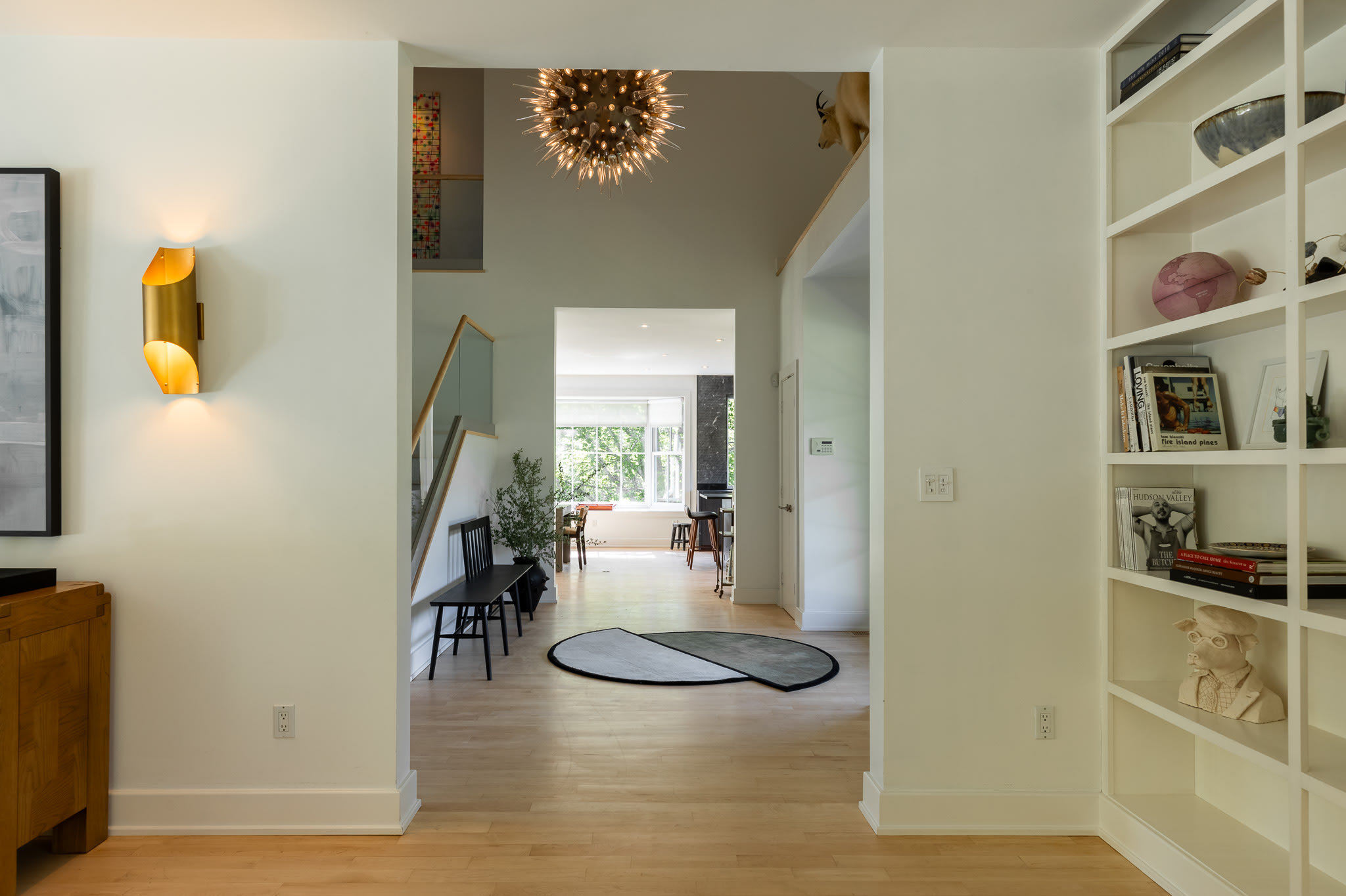 A bright hallway with light hardwood floors and white walls leads deeper into the house, revealing a staircase with a glass railing and a striking sputnik-style chandelier overhead. A round rug and a bench are visible in the foreground.