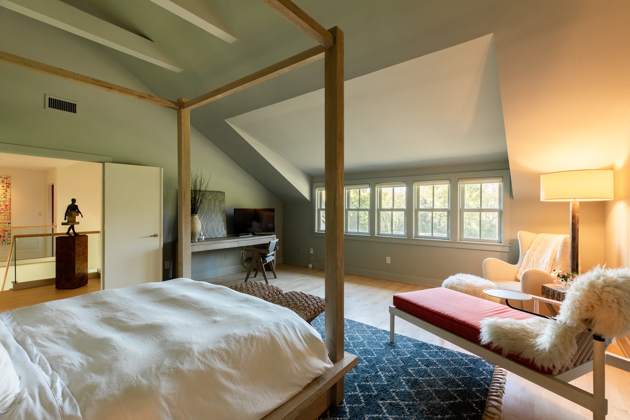 Spacious attic bedroom with a light wood canopy bed, light green walls, and a sitting area with a red bench and white armchair, all on light hardwood floors. A desk and TV are visible in the background.