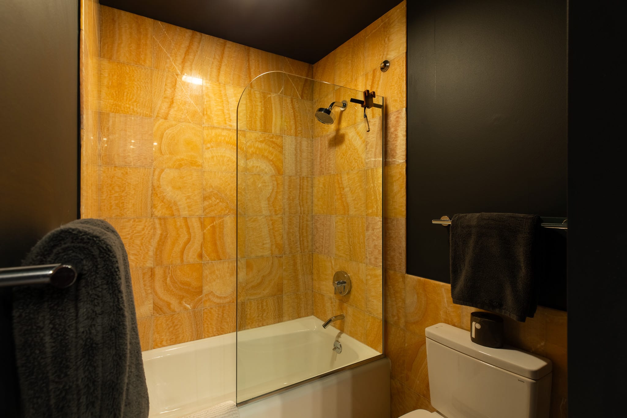 Bathroom featuring a bathtub with a glass shower screen, surrounded by vibrant orange-toned tiled walls, and a dark-painted wall with a toilet visible on the right.