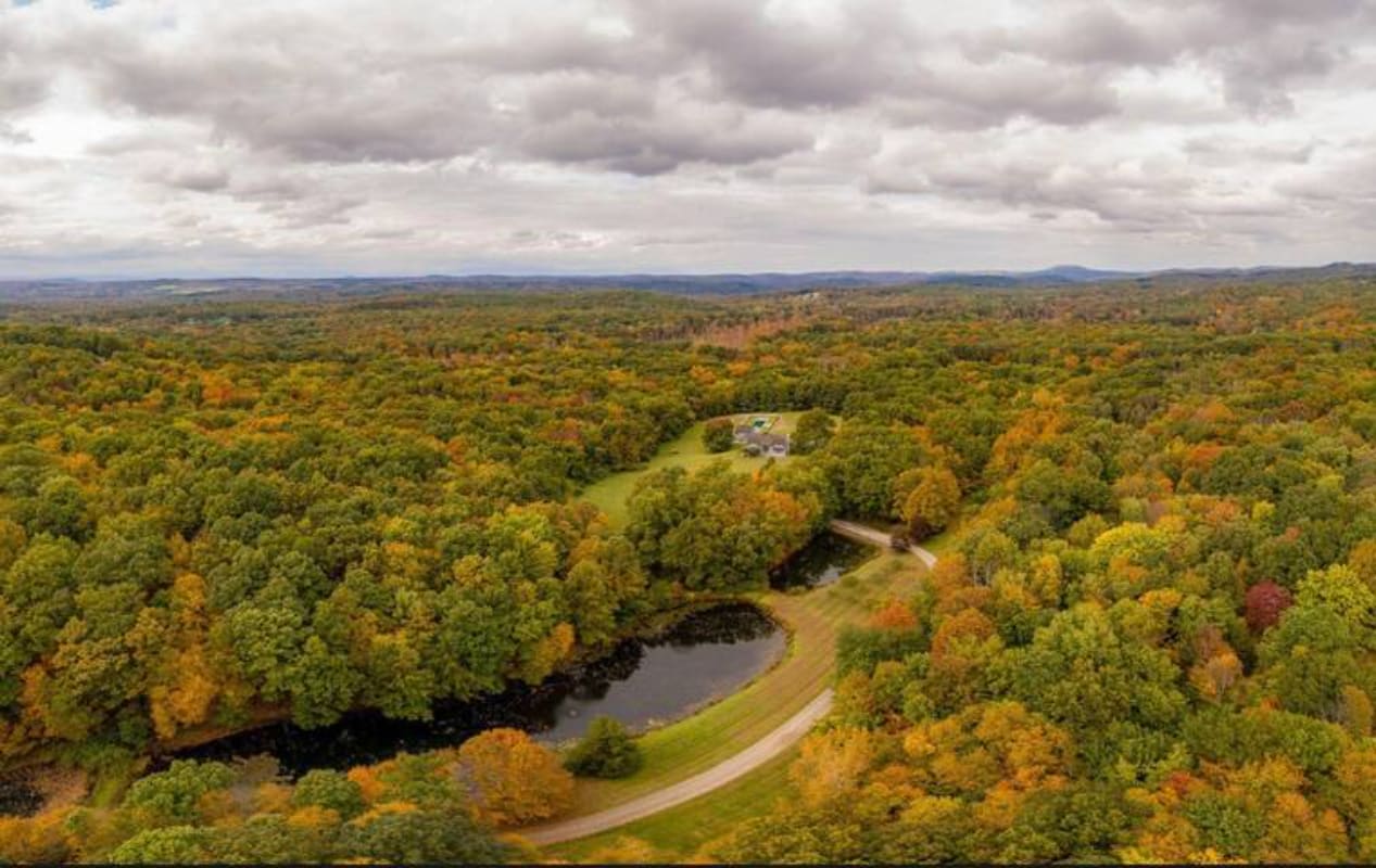 Expansive aerial view of a property nestled within a vast, colorful forest, showcasing autumn foliage. A winding road leads to the house, with a dark pond visible in the foreground, all under a cloudy sky.
