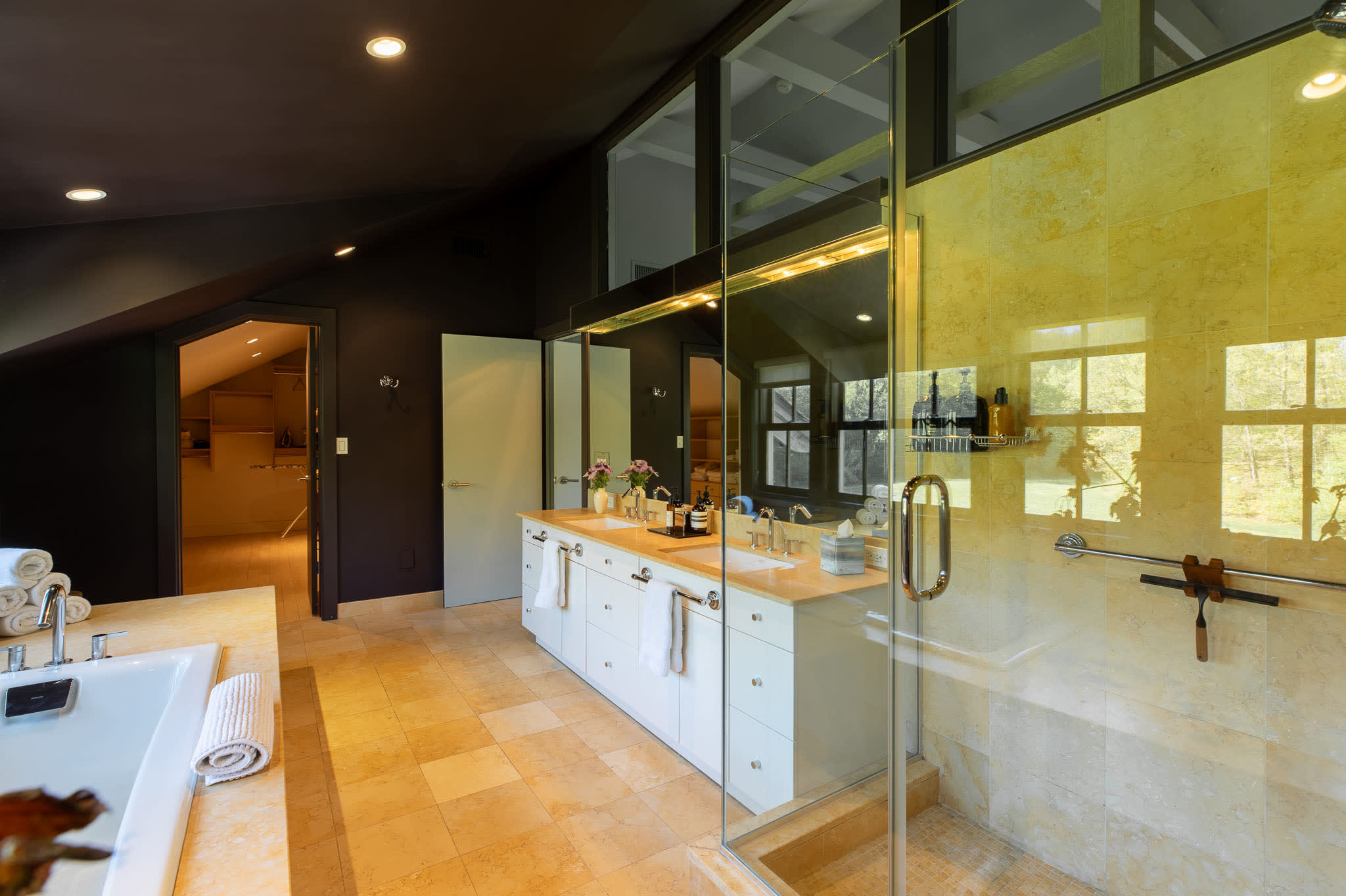 View of the glass-enclosed shower and double vanity with white cabinets and light countertops in a dark-walled bathroom, with a doorway leading to a walk-in closet.