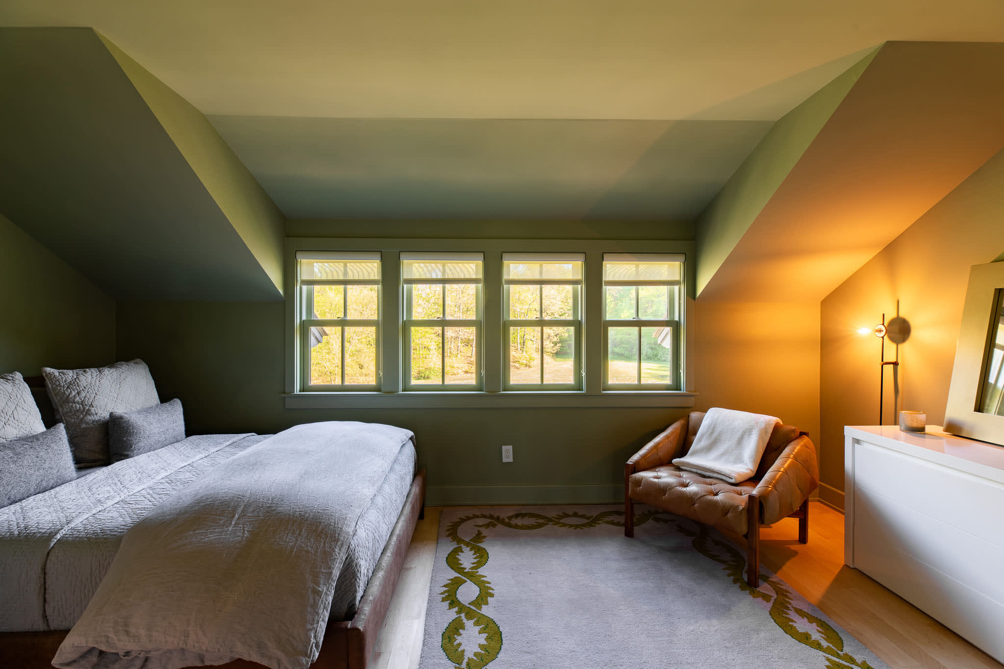 View of the olive green bedroom showing a comfortable brown leather armchair, a white dresser, and a floor lamp, with natural light streaming through the windows.