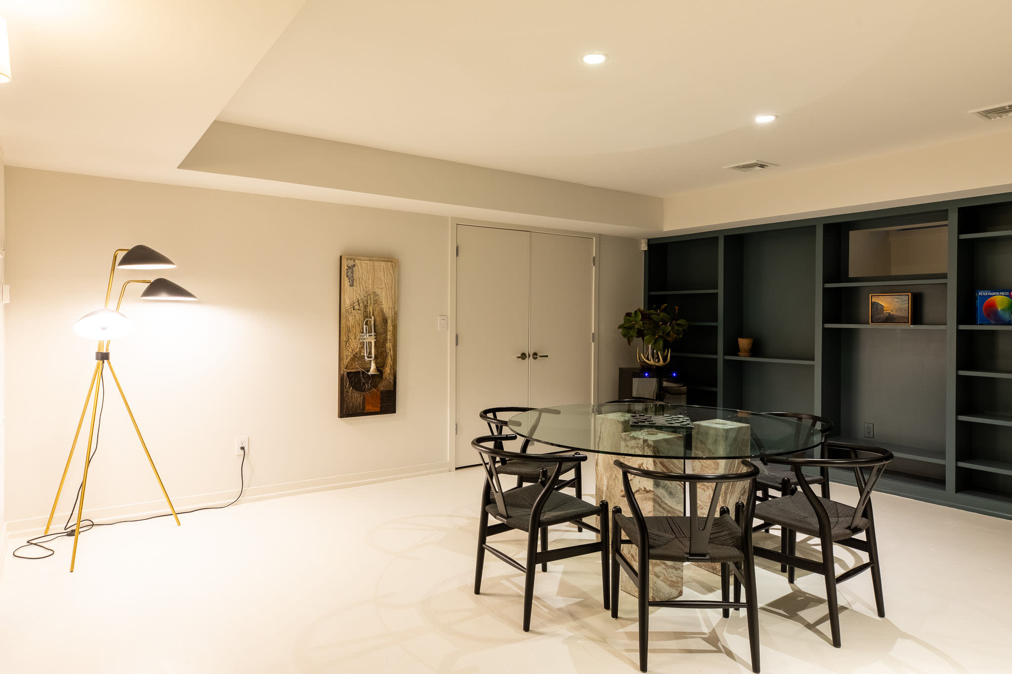 Elegant dining area featuring a round glass-top table with a unique stone base and six black wooden chairs, set against a backdrop of dark teal built-in bookshelves. A modern tripod floor lamp provides additional lighting.