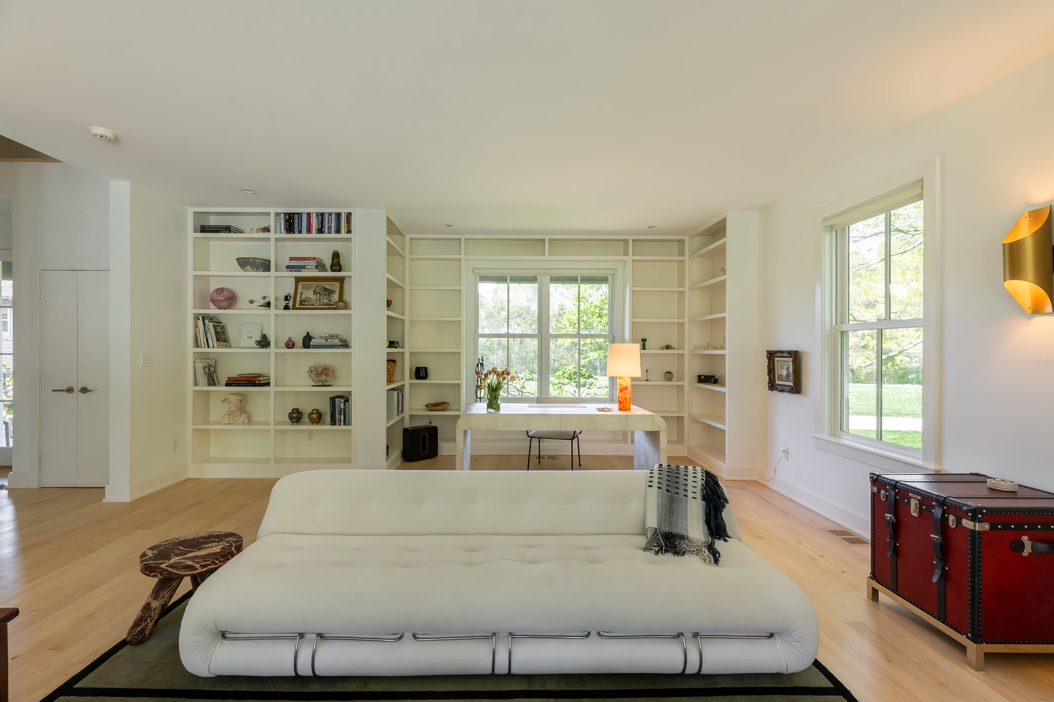 A wide shot of the living room showcases extensive white built-in bookshelves spanning the back wall, framing a large window and a white desk. A long white sofa sits on a dark rug, with light hardwood floors and white walls throughout.