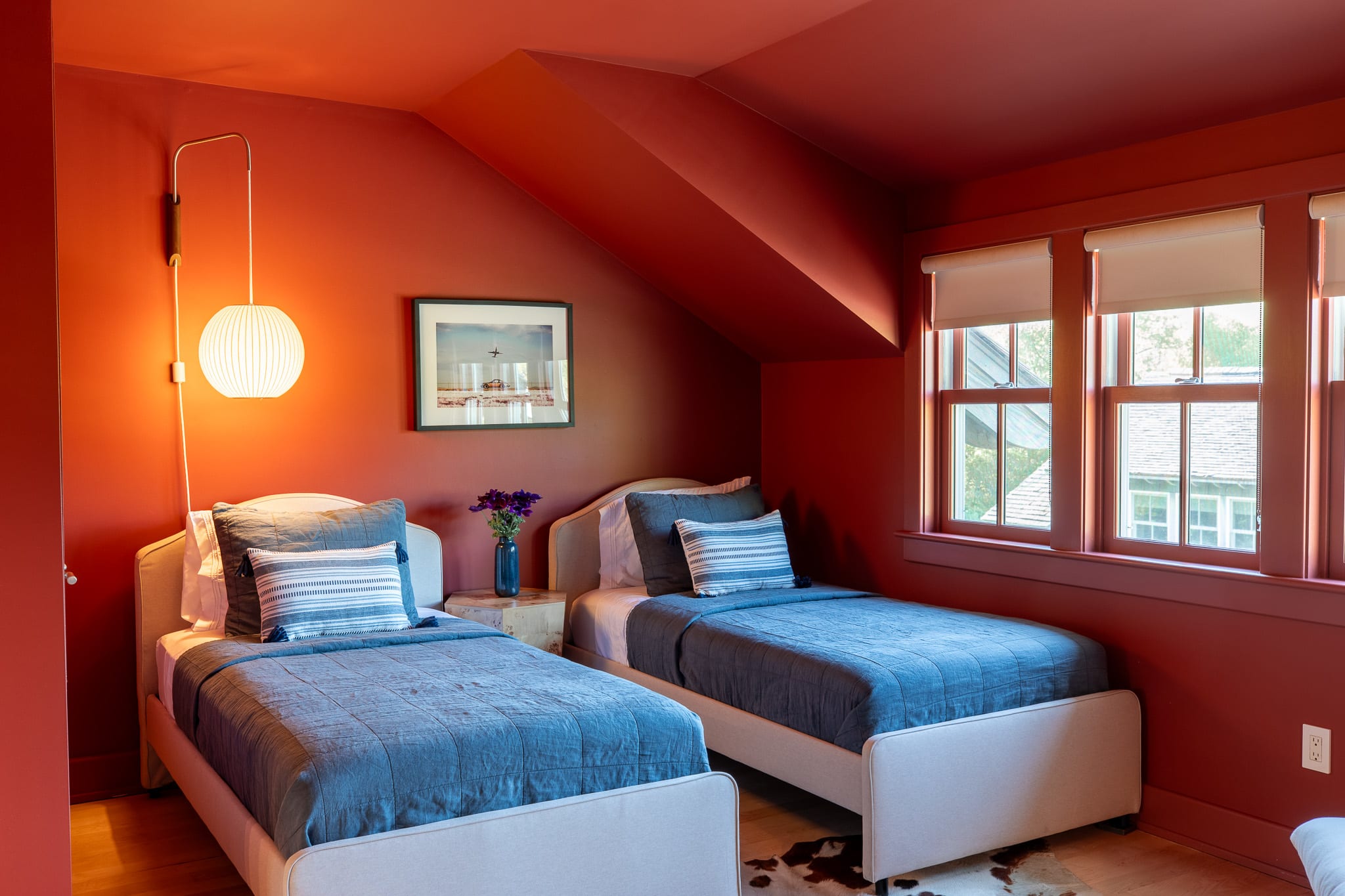 Cozy bedroom with two twin beds featuring blue bedding and striped pillows, set against striking red walls and a sloped ceiling, with a modern wall lamp and framed art.