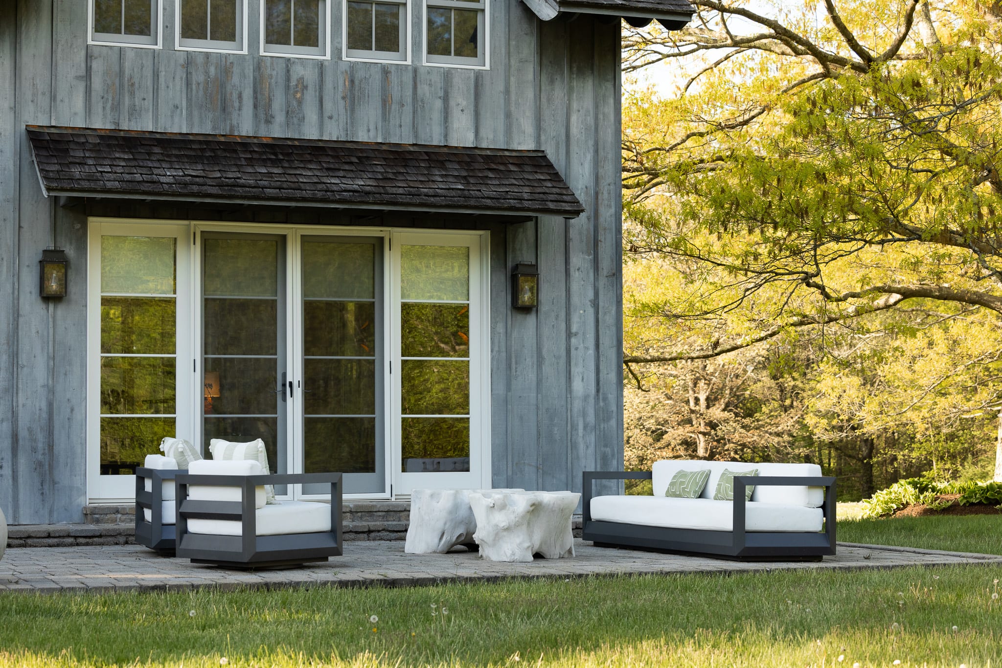 An expansive outdoor patio area with modern seating, including two armchairs and a sofa with white cushions and green patterned pillows, is arranged around organic-shaped white coffee tables. The patio is made of light pavers and borders a lush green lawn with mature trees in the background.