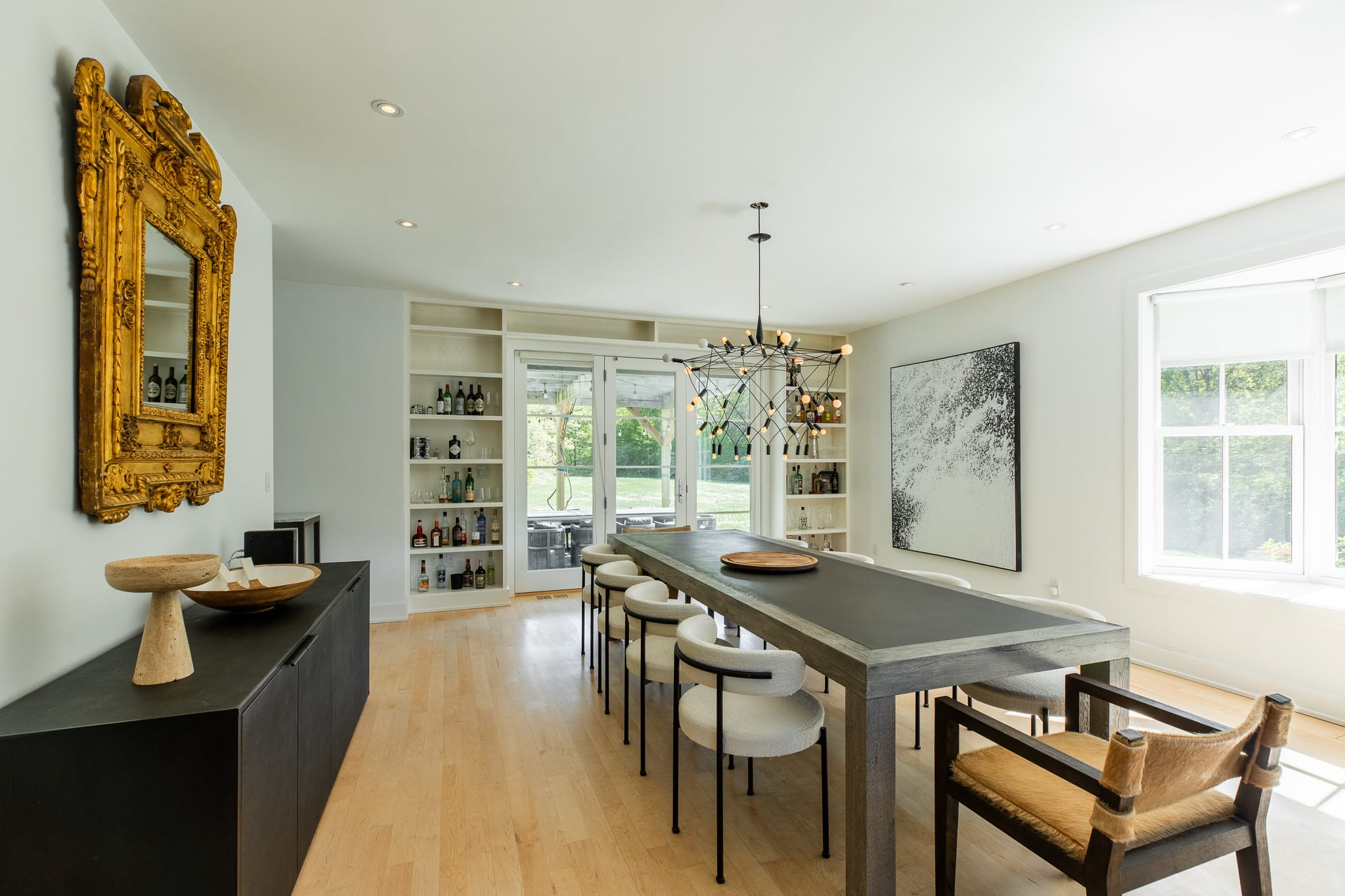 Wide view of the dining area with a long table and chairs, a large ornate gold mirror, and a dark credenza. Built-in shelves with bottles line the wall next to French doors.