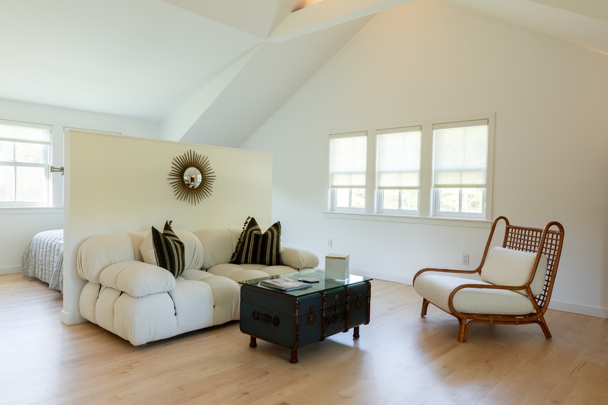 Spacious living area featuring a white sofa with striped pillows, a dark trunk coffee table, and a rattan armchair, with a partial view of a bed behind a white partition wall.