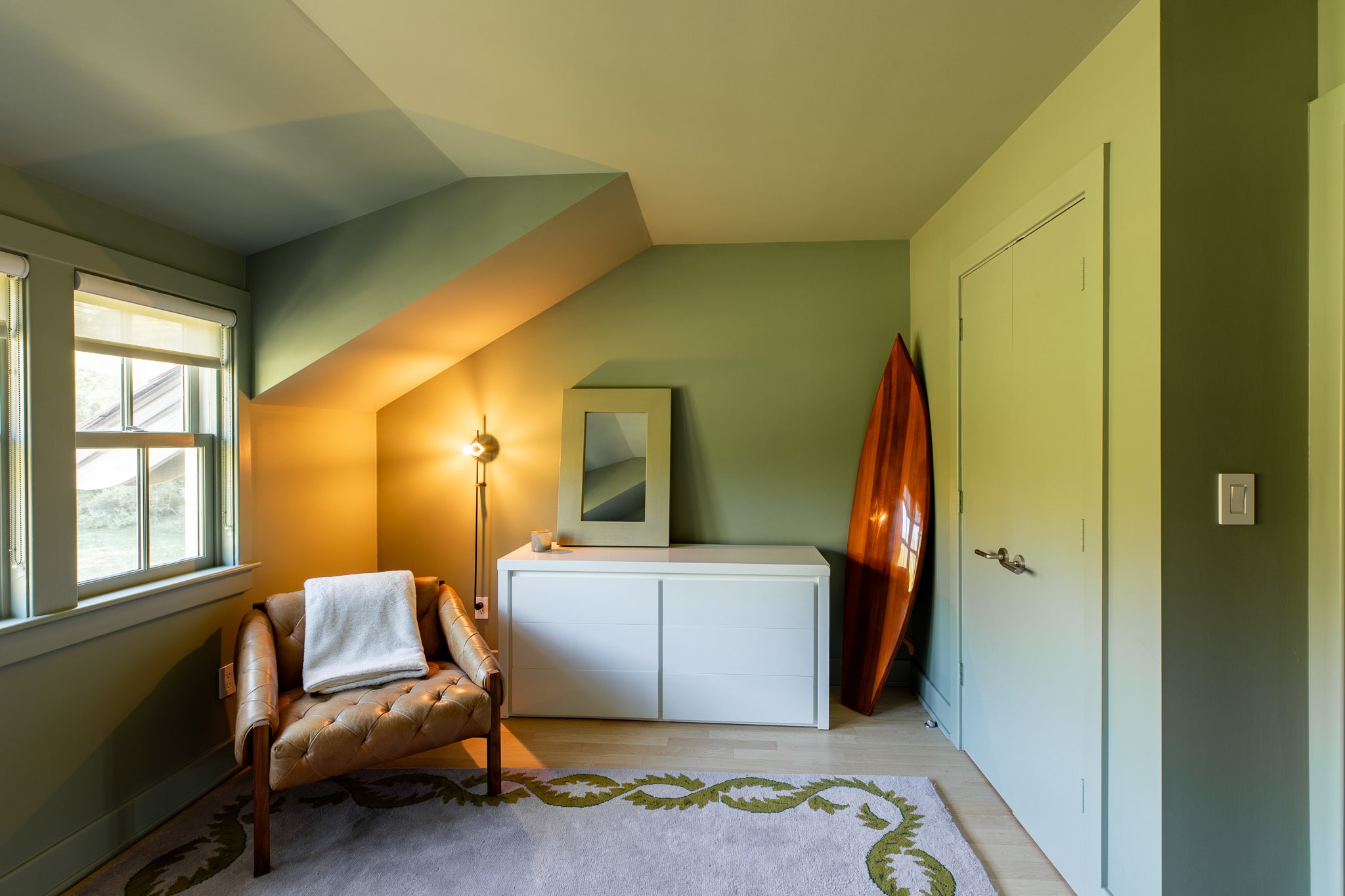 Corner of the olive green bedroom featuring a white dresser with a mirror, a floor lamp, and a decorative wooden surfboard leaning against the wall next to a closed door.