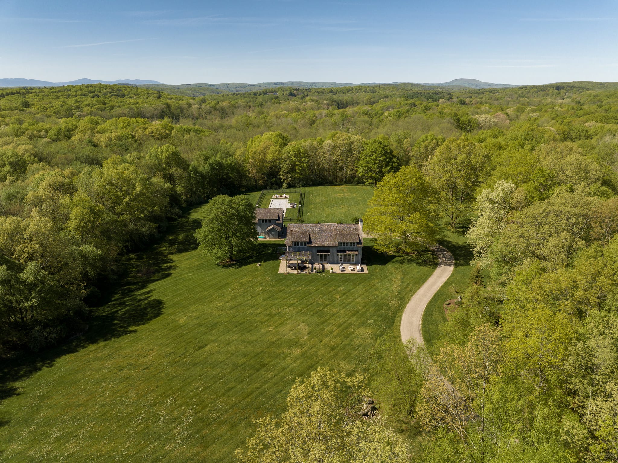 An aerial view showcases a large gray house with a light-colored driveway winding through expansive green lawns, surrounded by dense, vibrant green forests under a clear blue sky. Distant mountains are visible on the horizon.