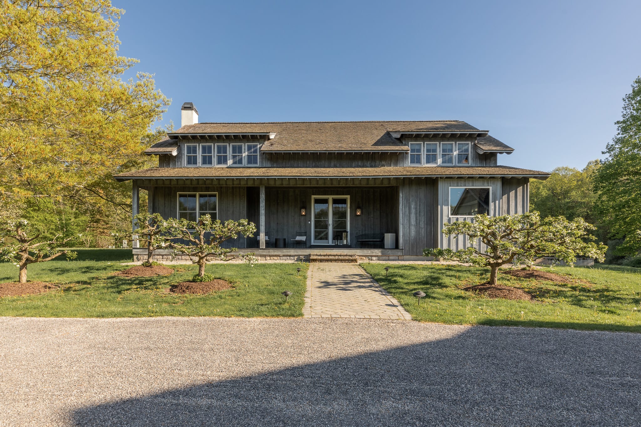 The front exterior of a charming two-story house with weathered gray wooden siding and a brown shingle roof, featuring multiple windows and a central entrance with glass doors. A paved walkway leads to the house, flanked by manicured small trees and a gravel driveway in the foreground.