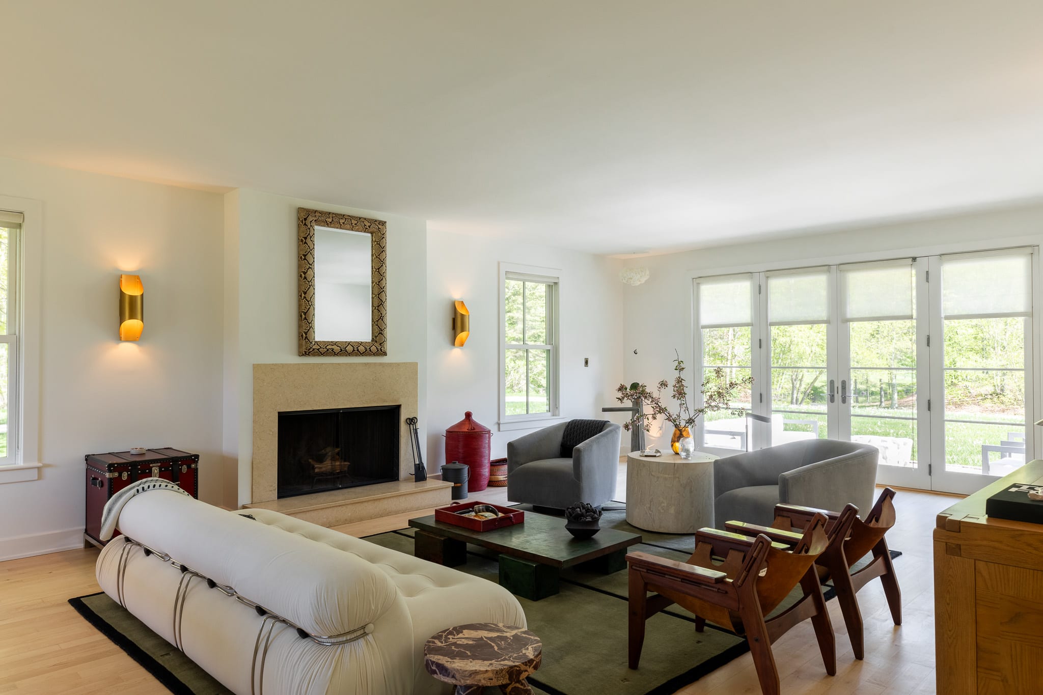 A spacious living room features light hardwood floors, white walls, and a stone fireplace with a decorative mirror above. A long white sofa, two gray armchairs, and a dark coffee table are arranged on a green rug, with French doors providing outdoor access.