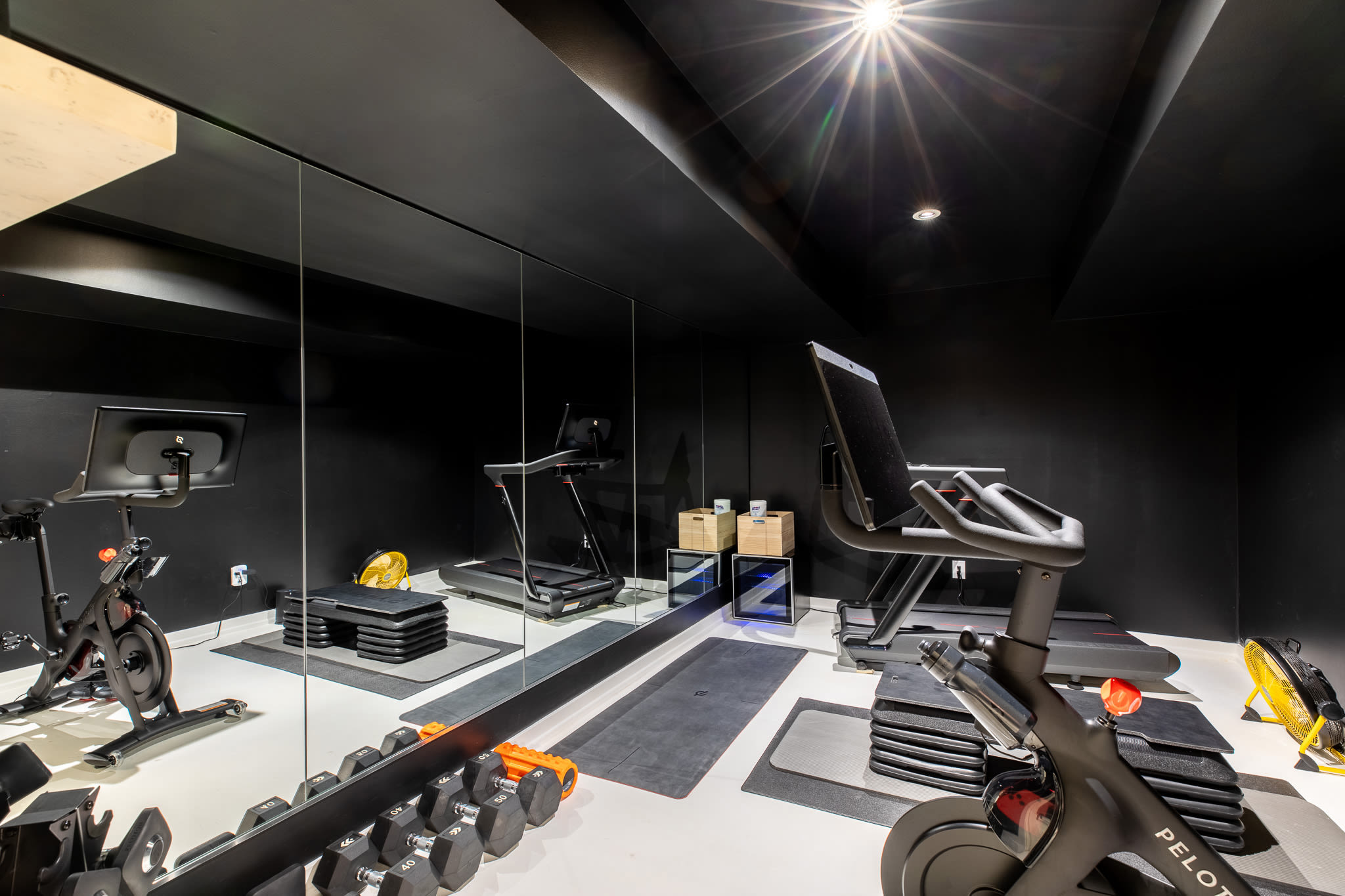 Modern home gym with sleek black walls and ceiling, a bright white floor, and a large mirrored wall reflecting exercise equipment like Peloton bikes, a treadmill, and dumbbells. Recessed lighting illuminates the space.