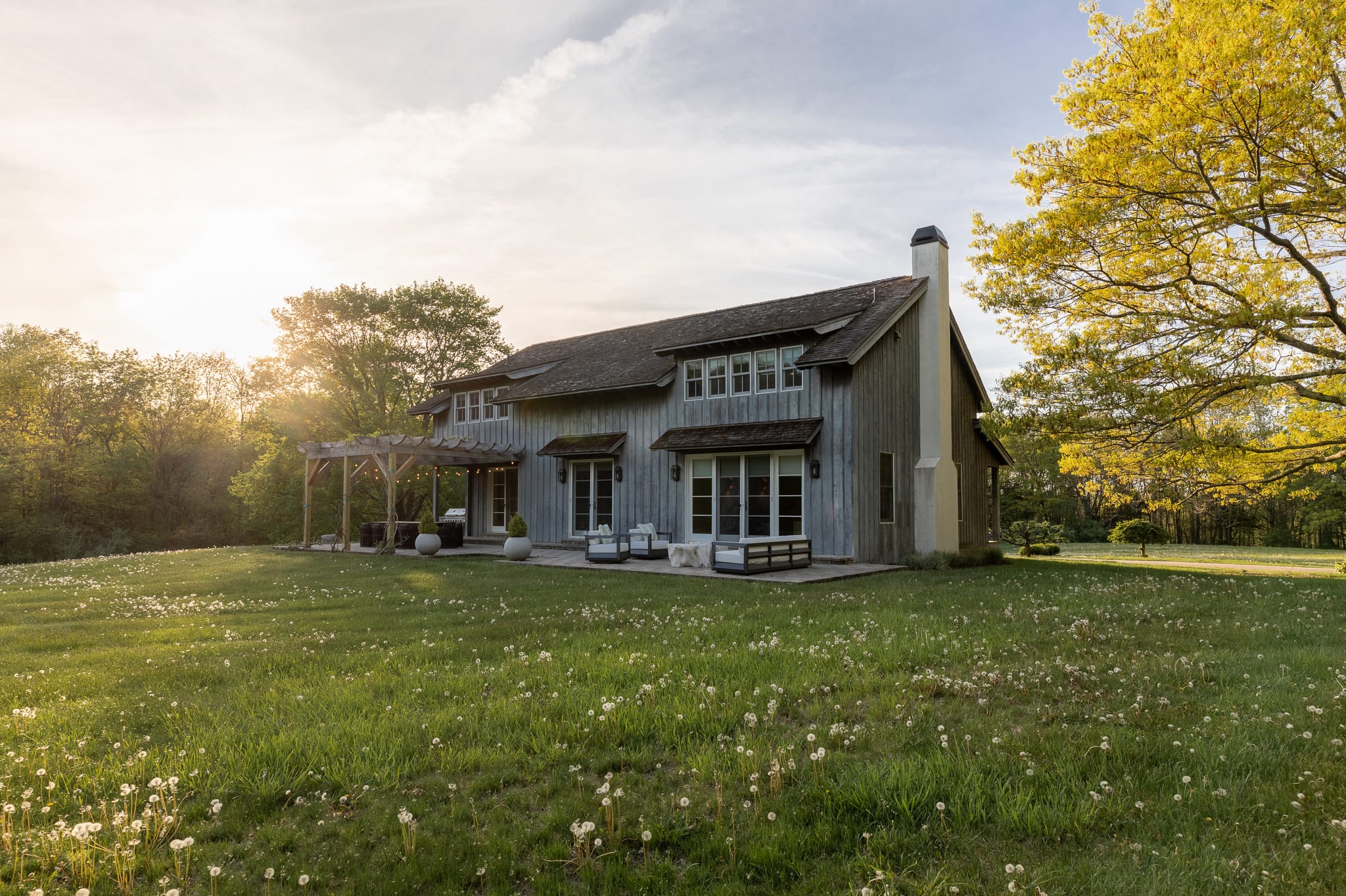 Charming rustic house with gray wooden siding and a shingled roof, featuring multiple windows and a covered patio area with outdoor seating. The house is surrounded by a lush green lawn dotted with dandelions, under a bright, sunny sky.