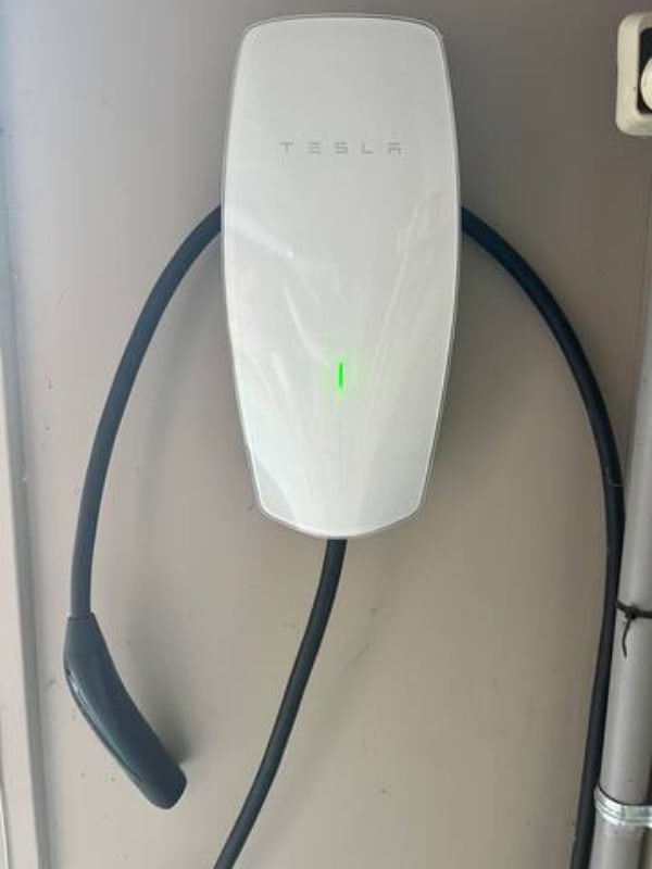 A white Tesla wall connector is mounted on a light-colored wall, with its charging cable neatly coiled around it. A green indicator light on the unit suggests it is powered on and ready for use.