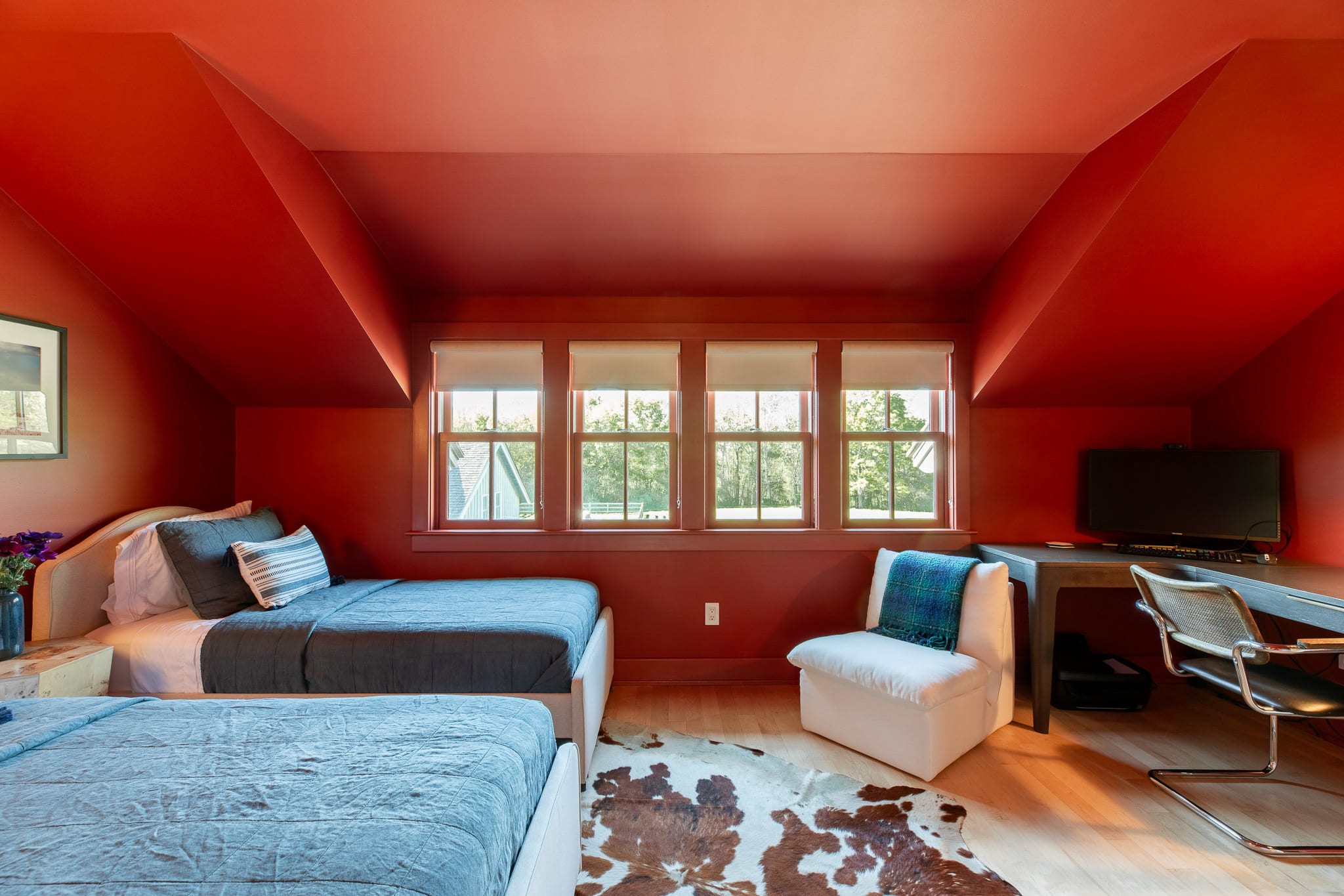 Wide view of a vibrant red-walled bedroom with two twin beds, a white armchair, and a desk area with a TV, featuring multiple windows overlooking greenery and a cowhide rug on the floor.