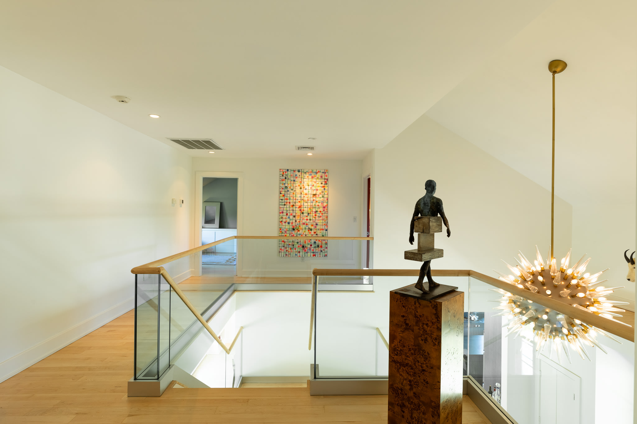 Bright upstairs landing with light hardwood floors and a glass railing overlooking the floor below. Modern art and a unique sputnik-style chandelier decorate the space.
