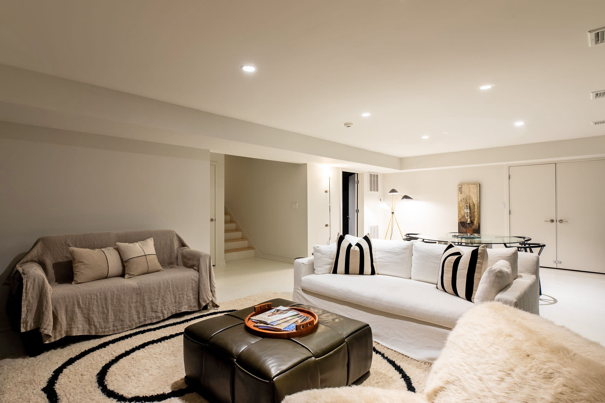 Expansive open-concept living area with a white slipcovered sofa, a dark leather ottoman, and a shaggy rug, flowing into a dining space with a glass-top table. Light-colored walls and recessed lighting create a bright, airy feel.