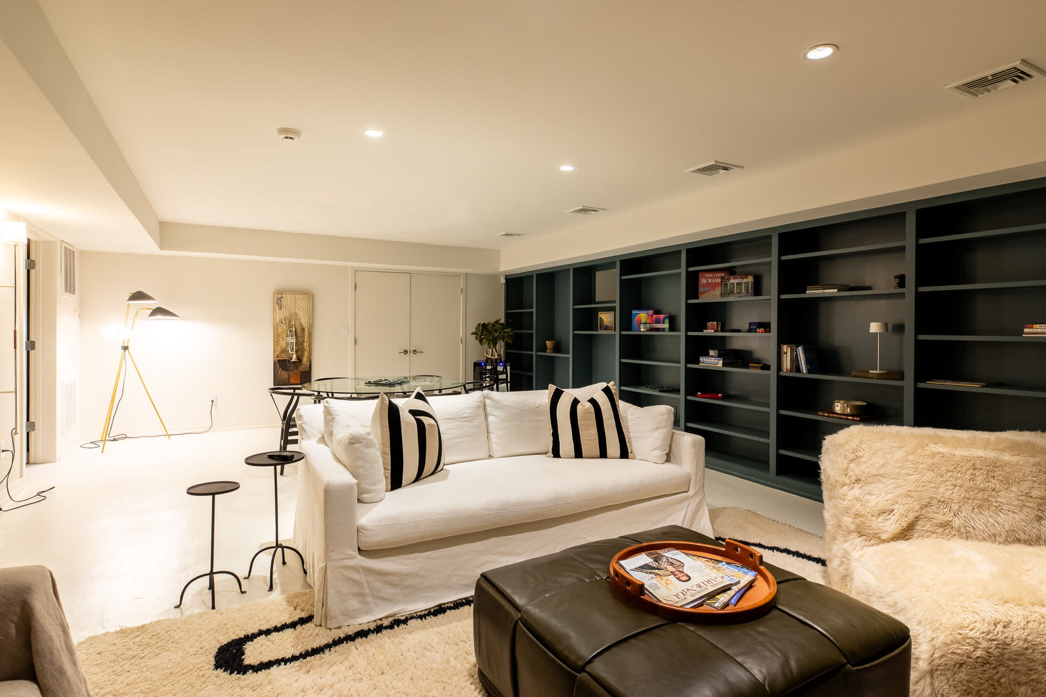 Cozy living area featuring a white slipcovered sofa with striped pillows, a dark leather ottoman, and a large built-in bookshelf unit in a dark teal color. A shaggy rug with a black and white pattern anchors the seating arrangement.