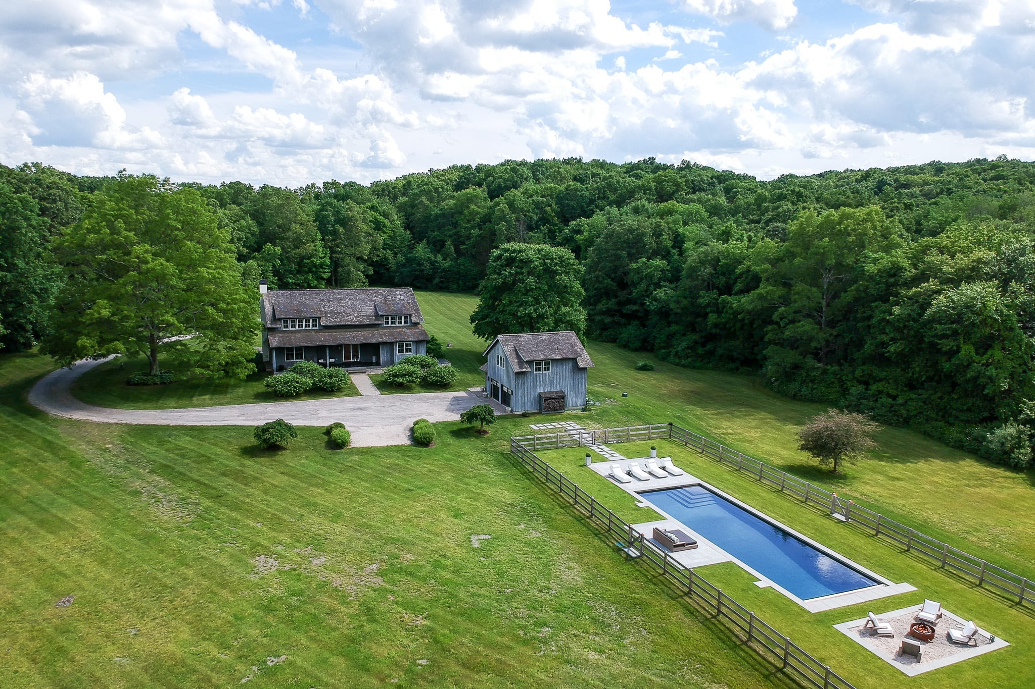 Stunning aerial view of a sprawling estate featuring a large main house, a separate guesthouse or barn, and a long rectangular swimming pool surrounded by a fence. The property is set amidst vast green lawns and dense woodlands under a partly cloudy sky.