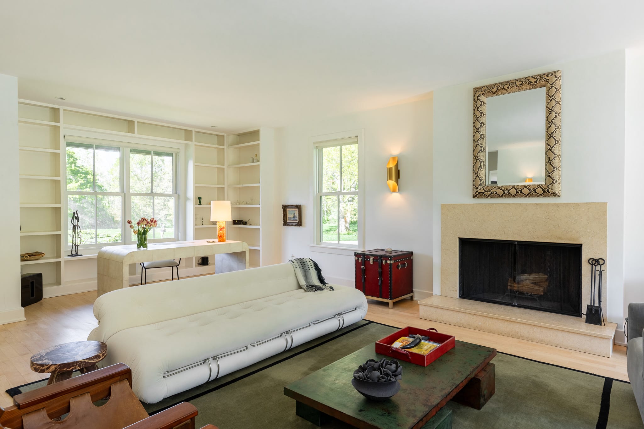 This living room view highlights a long white sofa facing a stone fireplace, with light hardwood floors and white walls. Built-in bookshelves line the far wall, framing a window and a desk area, while a red trunk adds a pop of color.