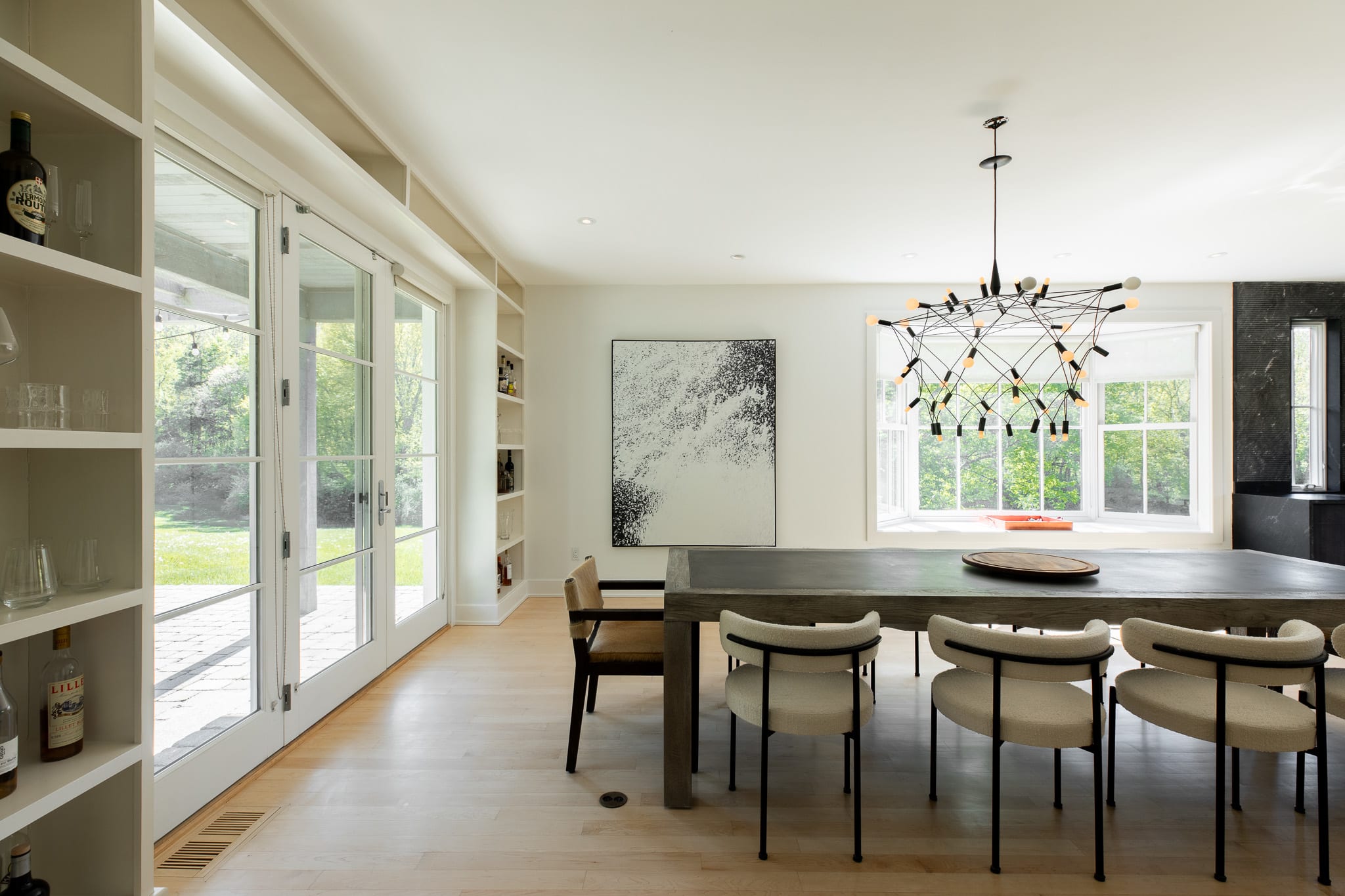 Dining area with a long dark wood table and modern chairs, featuring a large abstract painting on the wall and a contemporary chandelier. French doors lead to an outdoor patio, flanked by built-in shelves.