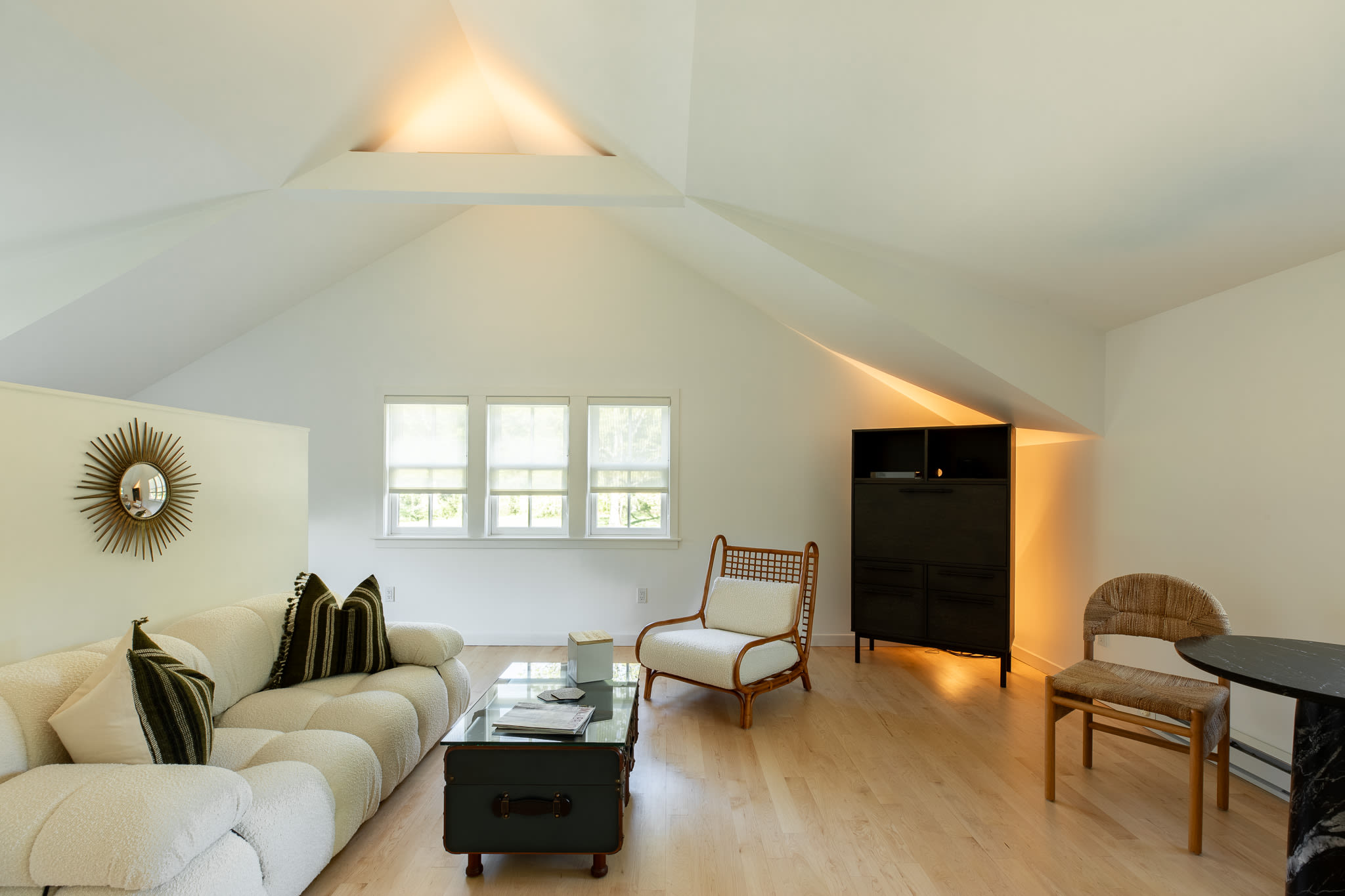 Bright living area with a white textured sofa, a vintage trunk coffee table, and a rattan armchair, set under a high vaulted ceiling with light wood floors and large windows.