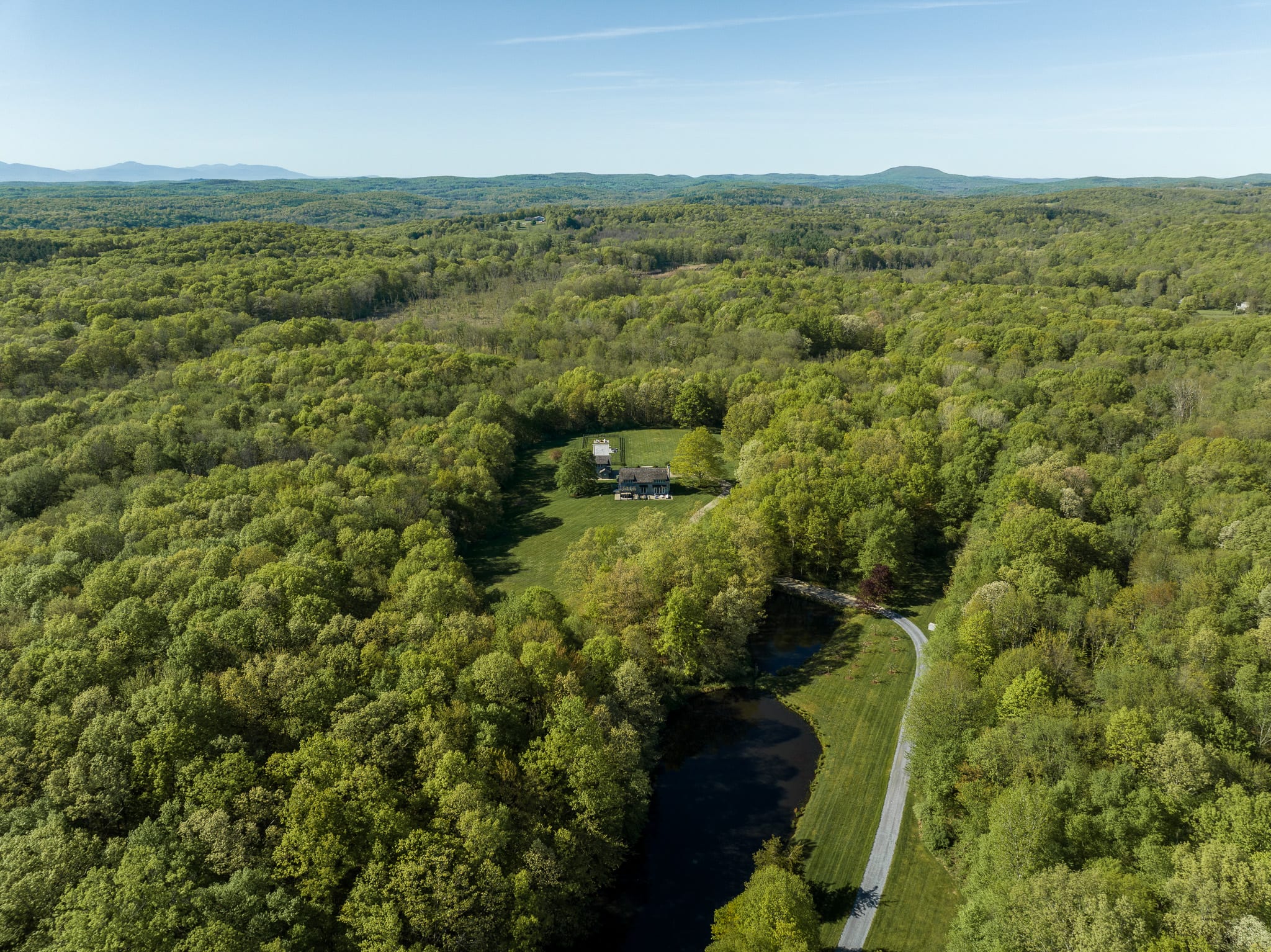 An expansive aerial view showcases a large property nestled within a vast, verdant forest under a clear blue sky. A winding driveway leads to a light-colored house, a dark pond, and an additional building, all surrounded by lush green trees and distant mountains.