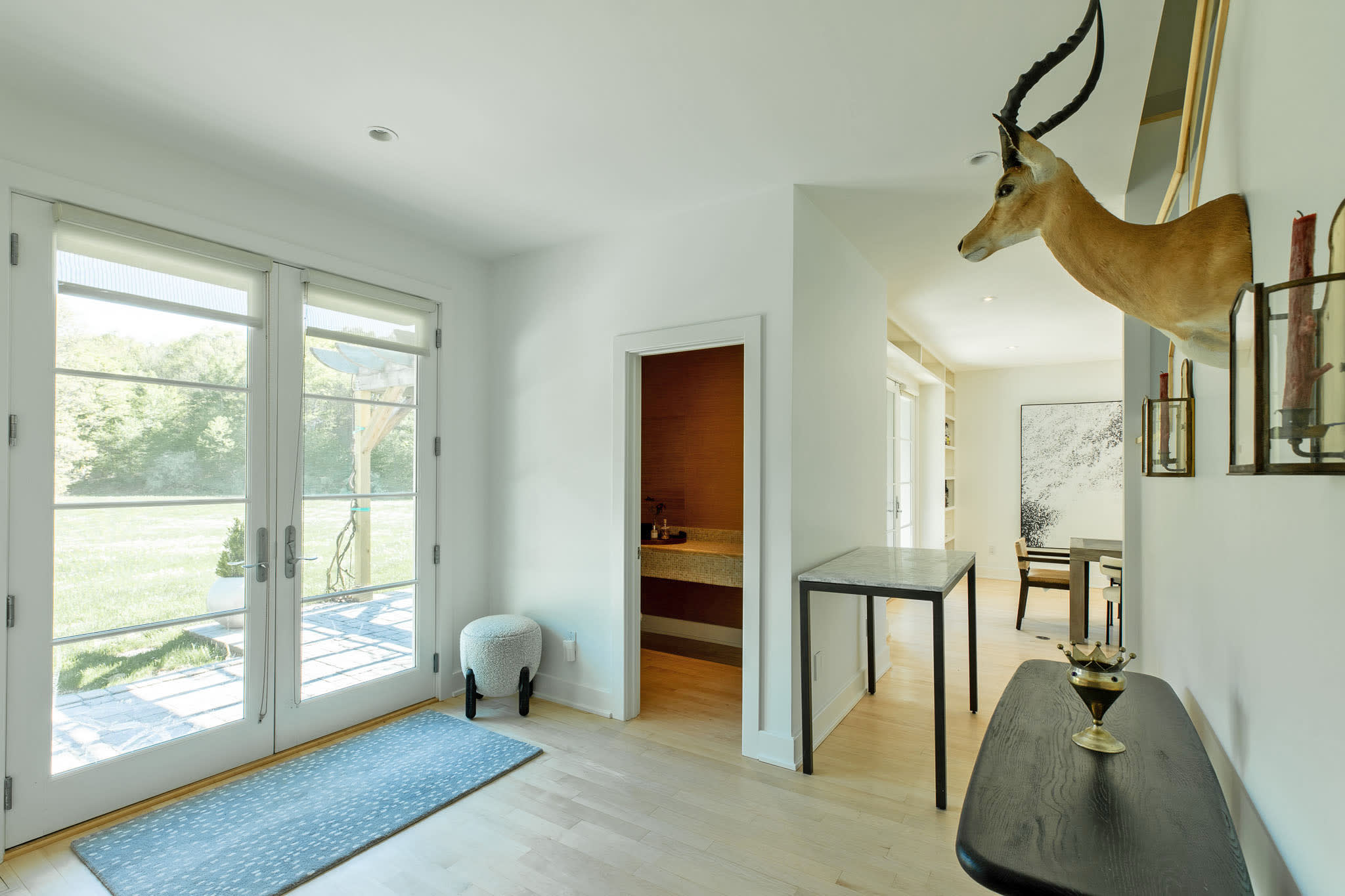 A bright entryway features light hardwood floors, white walls, and French doors leading to an outdoor patio. A taxidermy gazelle head is mounted on the wall, and a small powder room with a dark vanity is visible.