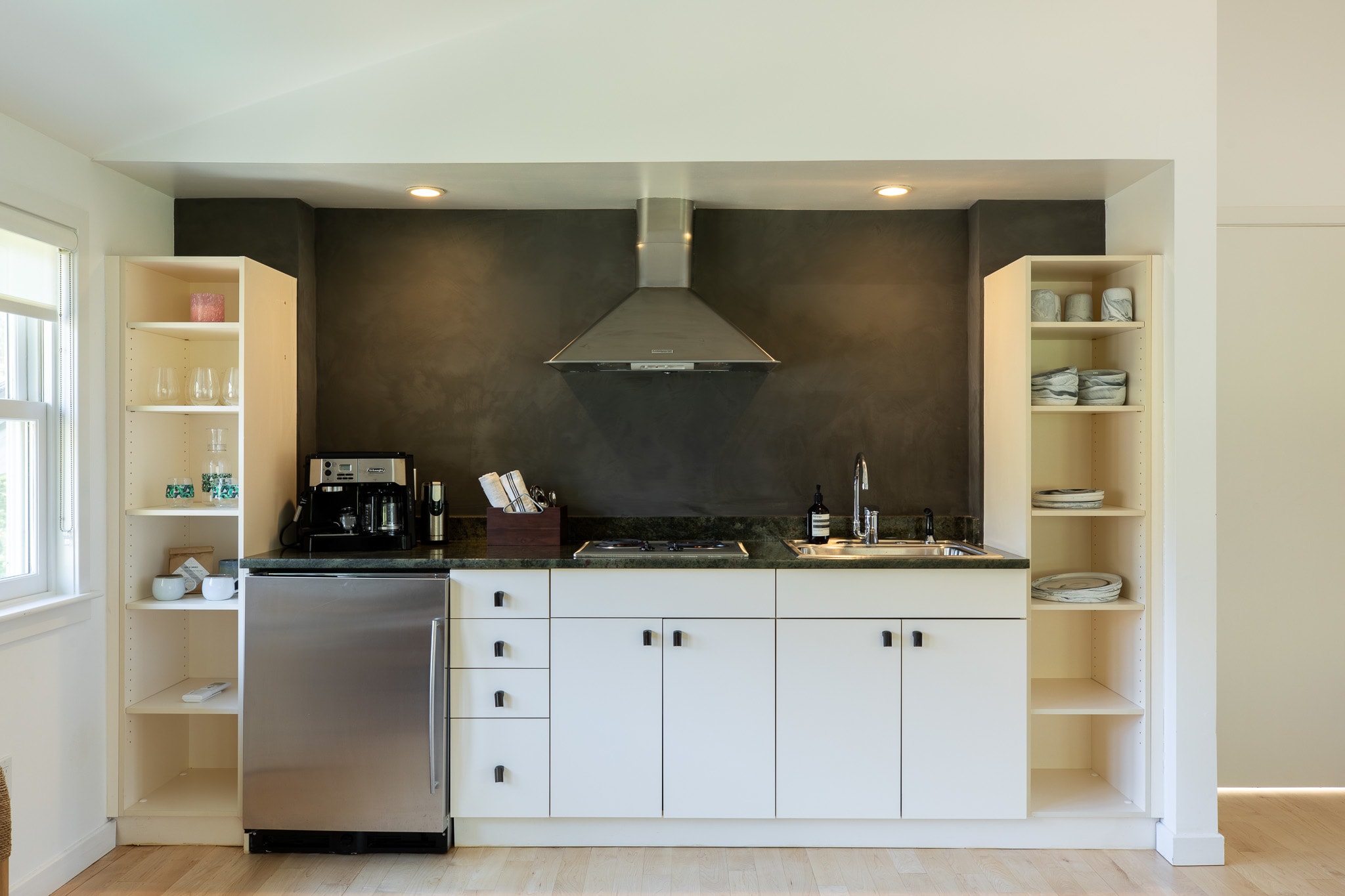 Compact kitchenette with white base cabinets, a dark countertop, stainless steel refrigerator, and a range hood, flanked by open shelving and set against a dark accent wall.