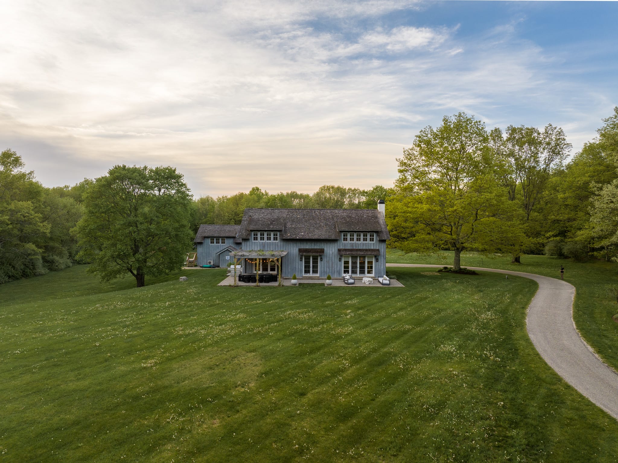 An aerial view captures the back of a light blue house with a dark roof, situated on a sprawling green lawn with a winding gravel driveway. Outdoor seating areas are visible on a patio, surrounded by mature trees and lush greenery under a partly cloudy sky.