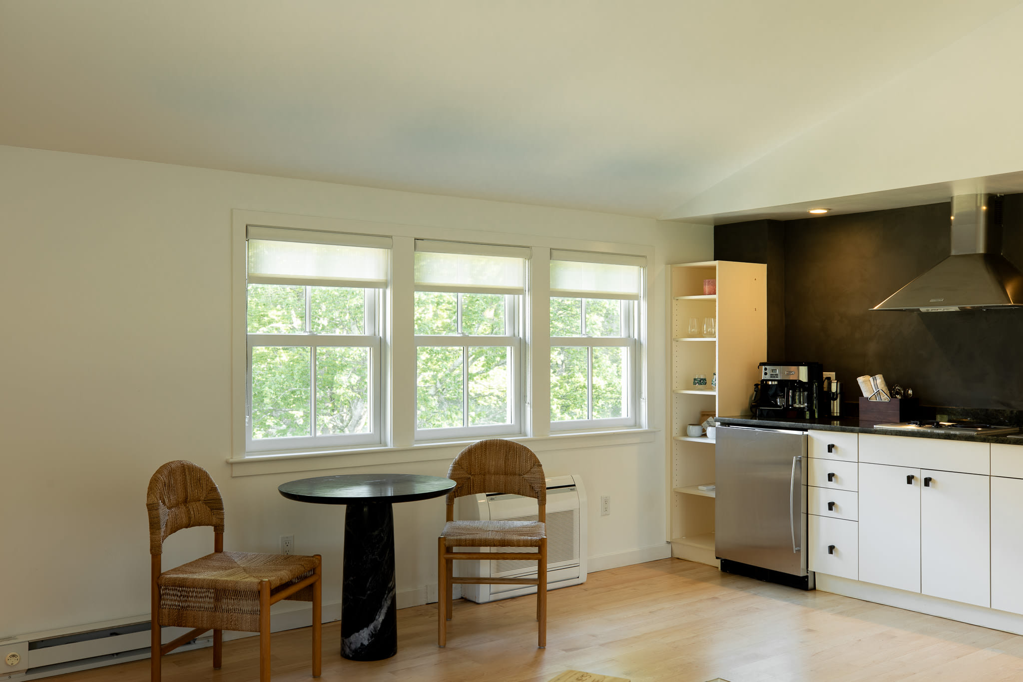 Bright kitchenette and dining area with white cabinets, a dark accent wall, and a small round table with two woven chairs, illuminated by natural light from multiple windows.
