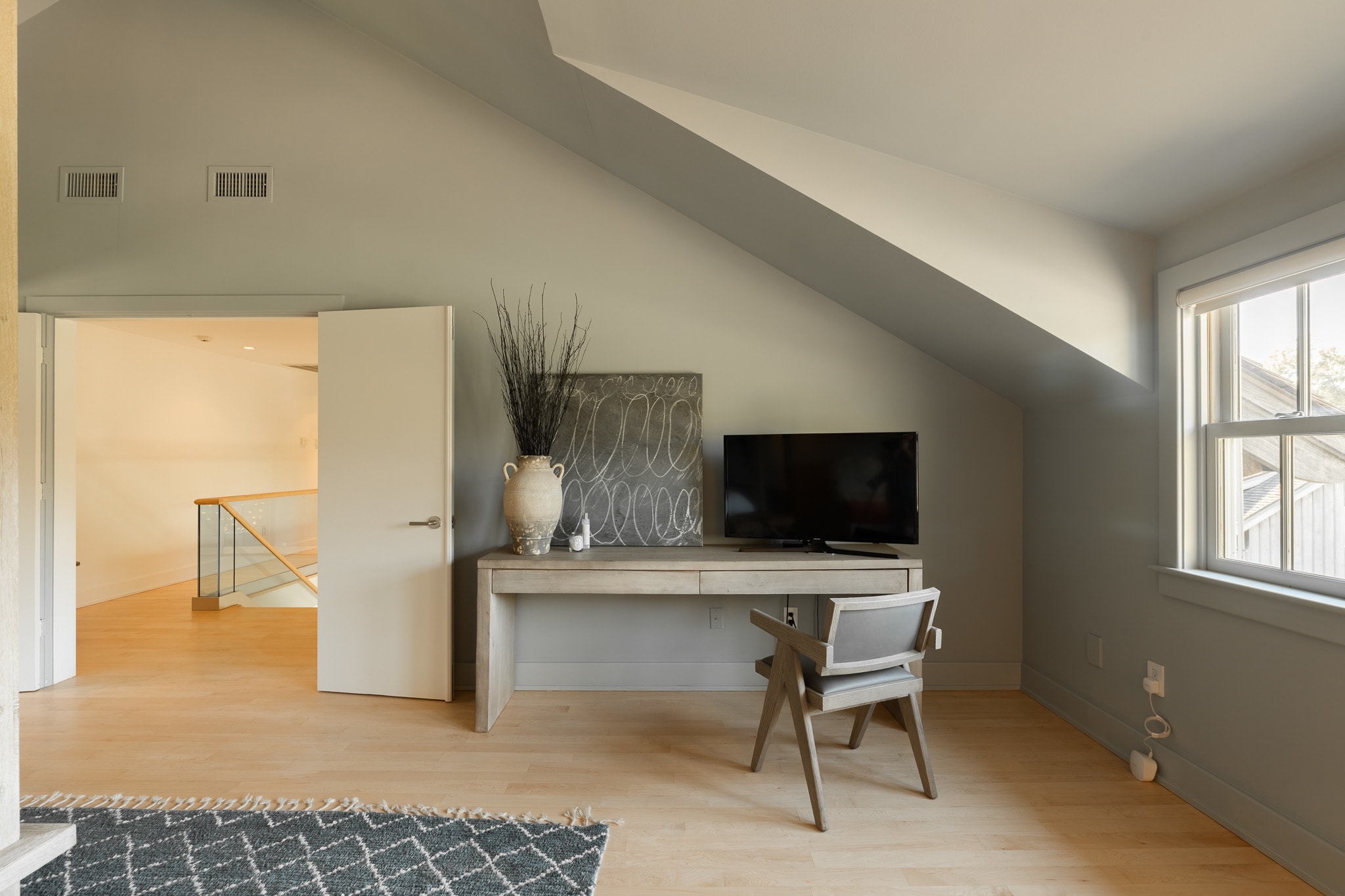 A light wood desk with a TV and a modern chair sits against a light green wall in an attic bedroom, featuring light hardwood floors and a doorway leading to a hallway.