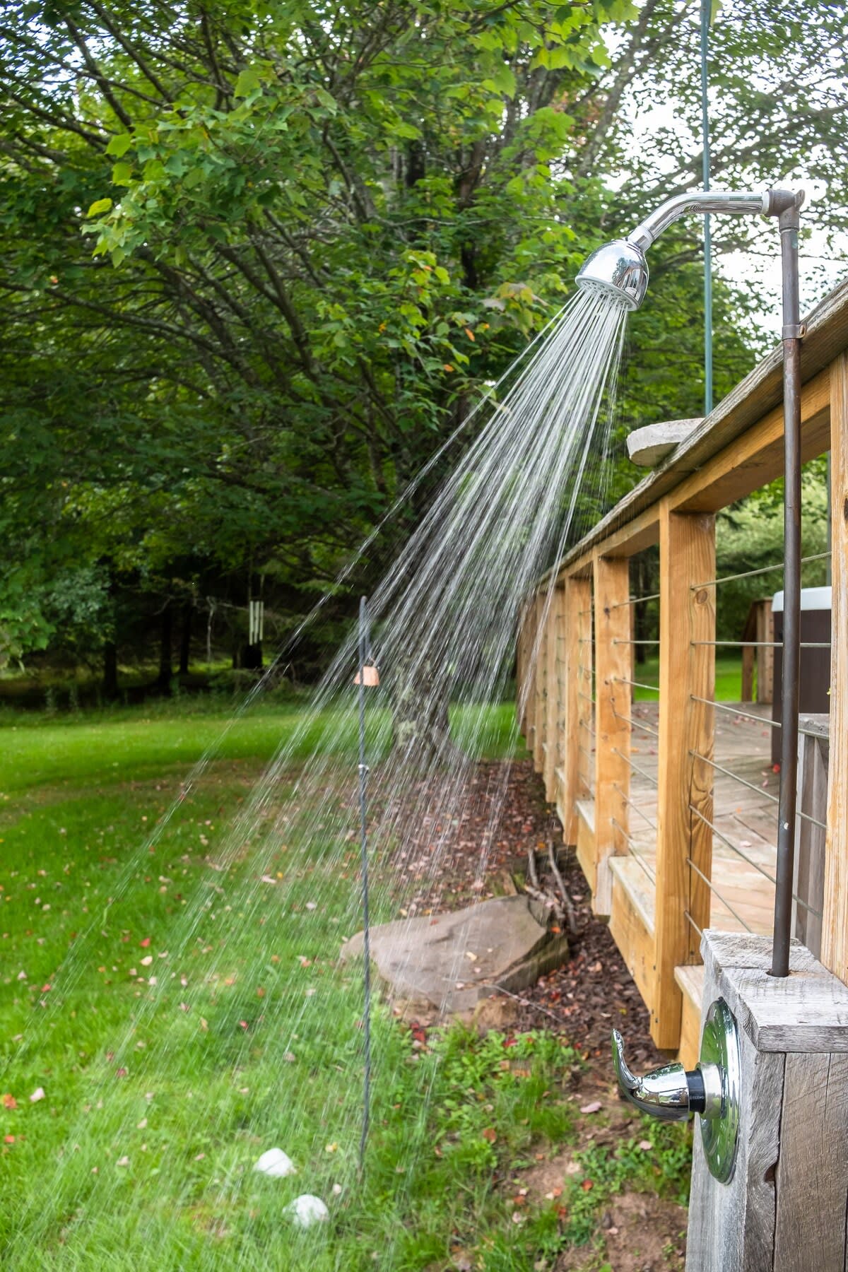 An outdoor shower with water spraying from the showerhead, set against a backdrop of a lush green lawn and dense trees. The shower fixture is mounted on a wooden post next to a wooden deck railing.