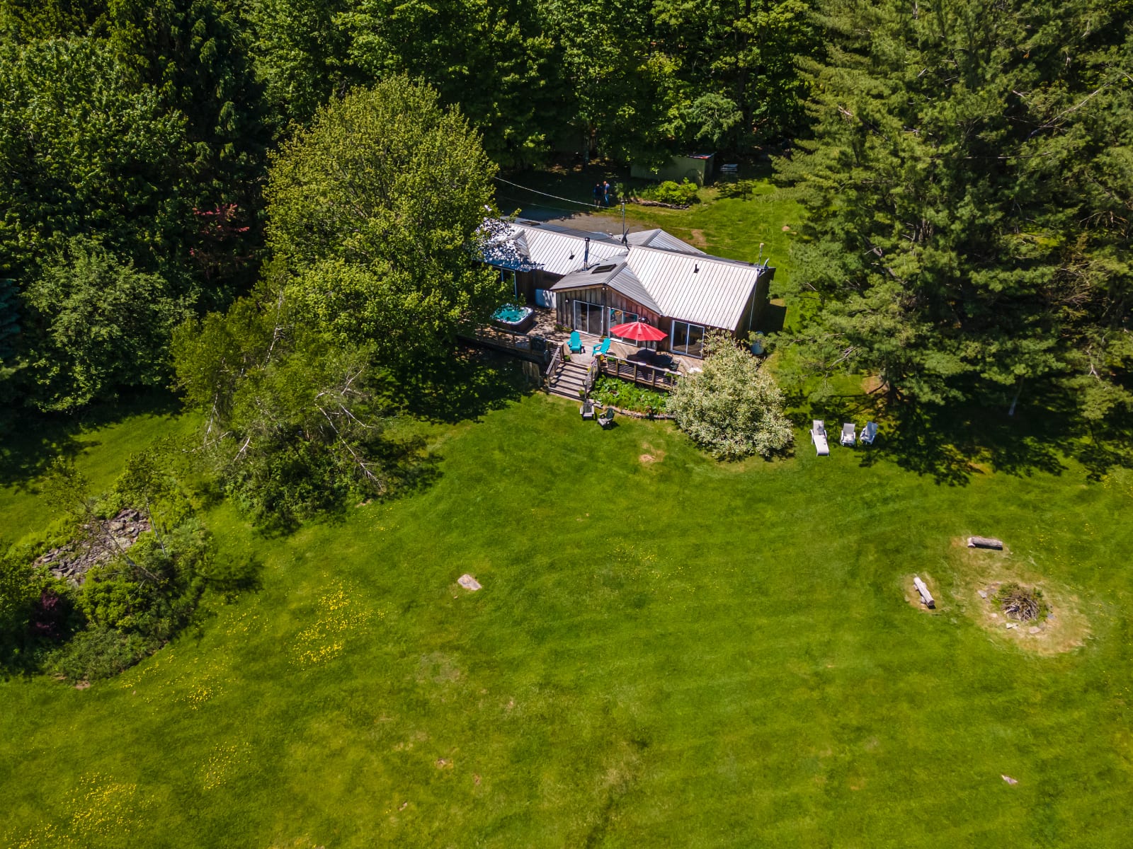 An aerial view of a rustic wooden cabin with a metal roof and a large deck, situated in a sprawling green lawn surrounded by dense trees, with a hot tub and outdoor seating visible on the deck.