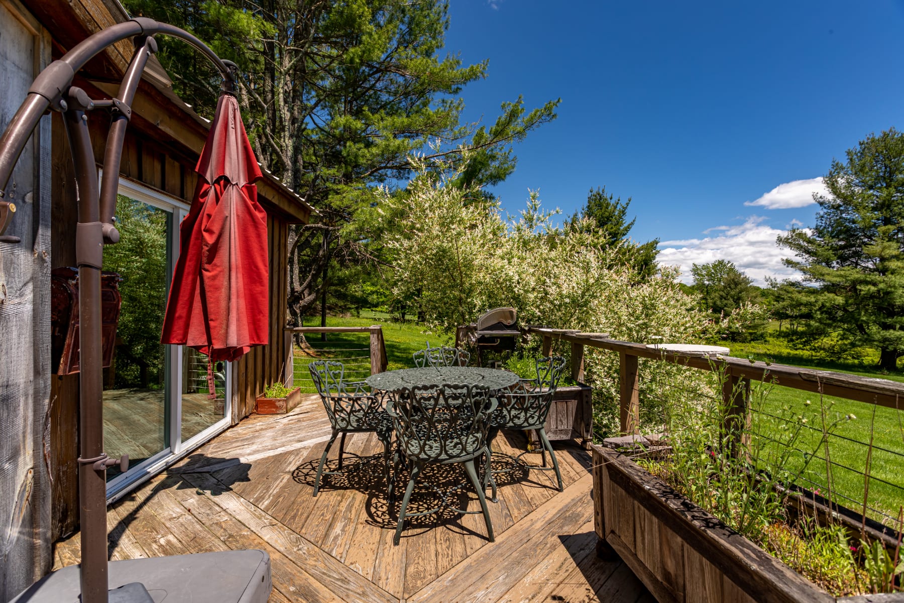 An outdoor wooden deck with a dark green metal dining set, including a table and four chairs, under a red patio umbrella. The deck overlooks a lush green yard and distant trees under a clear blue sky.