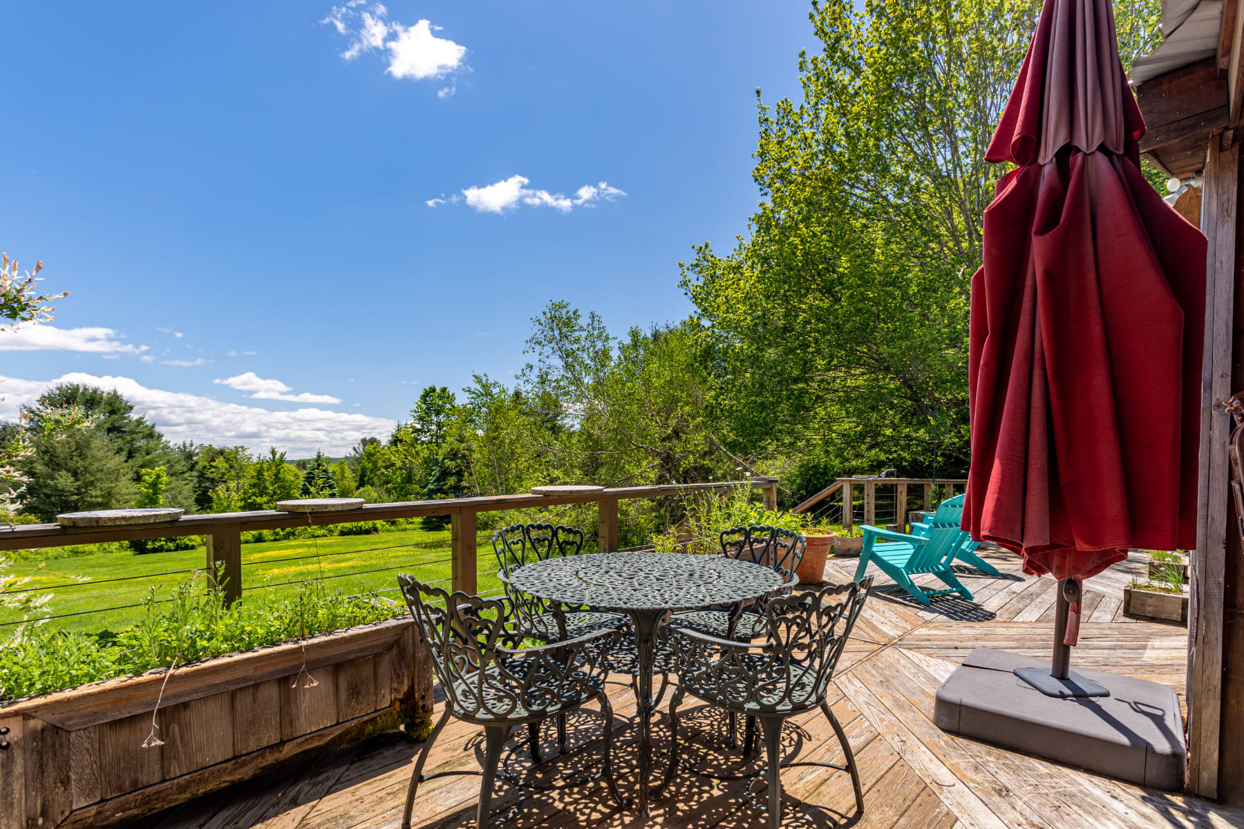 A spacious wooden deck features a round black metal outdoor dining set with chairs and a large red umbrella, offering a pleasant spot for al fresco dining amidst the surrounding green landscape and trees.